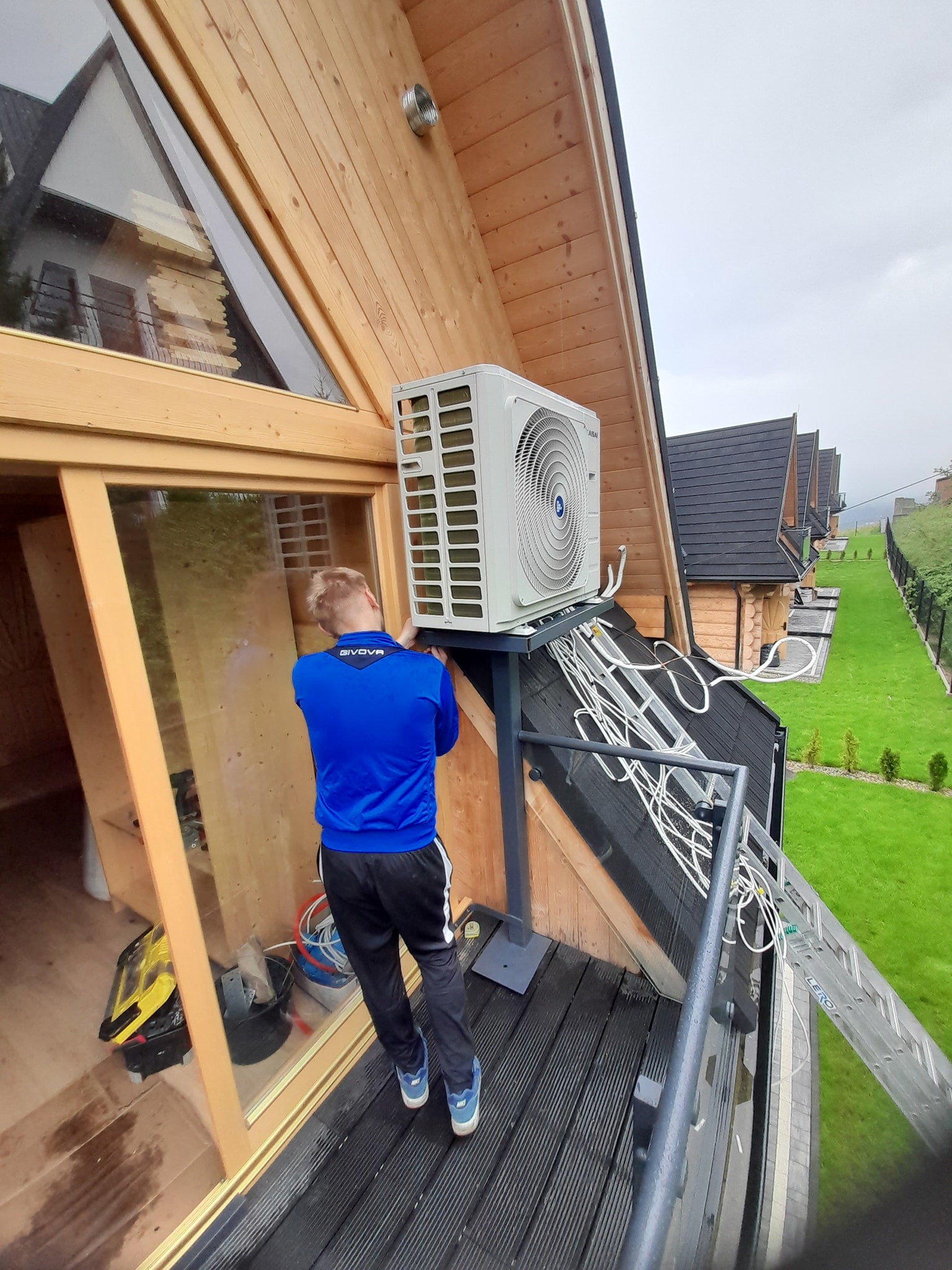 Montaż zewnętrznej jednostki klimatyzacji na wsporniku przy drewnianym domu w górach, widoczne przewody i drabina oparta o balkon.