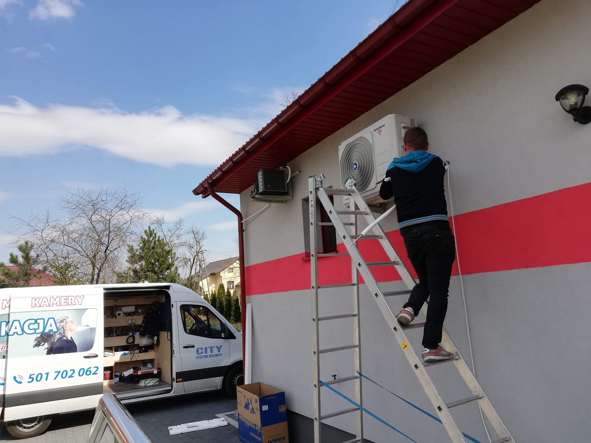Instalator na drabinie montuje klimatyzator marki Kaisai na elewacji domu z czerwonymi pasami, obok zaparkowany van z reklamą firmy instalacyjnej.