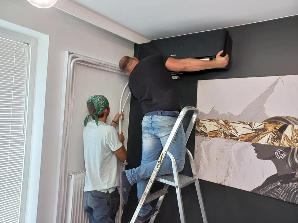 Dwóch monterów instaluje czarną jednostkę klimatyzacji w pomieszczeniu z ciemną ścianą i obrazem kobiety w złotej masce. Jeden monter na drabinie przykłada urządzenie do ściany, drugi prowadzi...