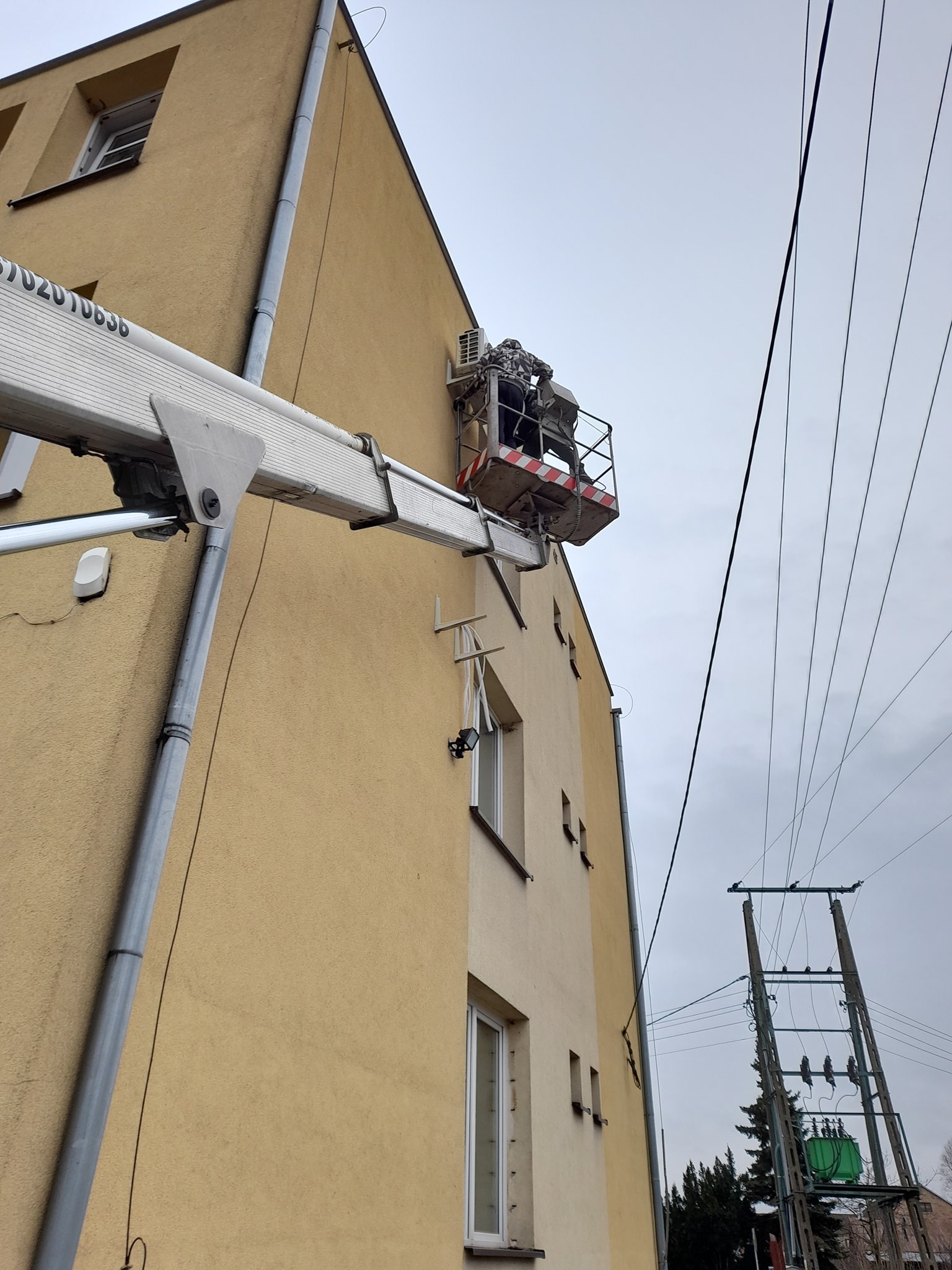 Pracownik w podnośniku koszowym montuje jednostkę zewnętrzną klimatyzacji na elewacji budynku mieszkalnego, widoczne przewody elektryczne i rynna spustowa.
