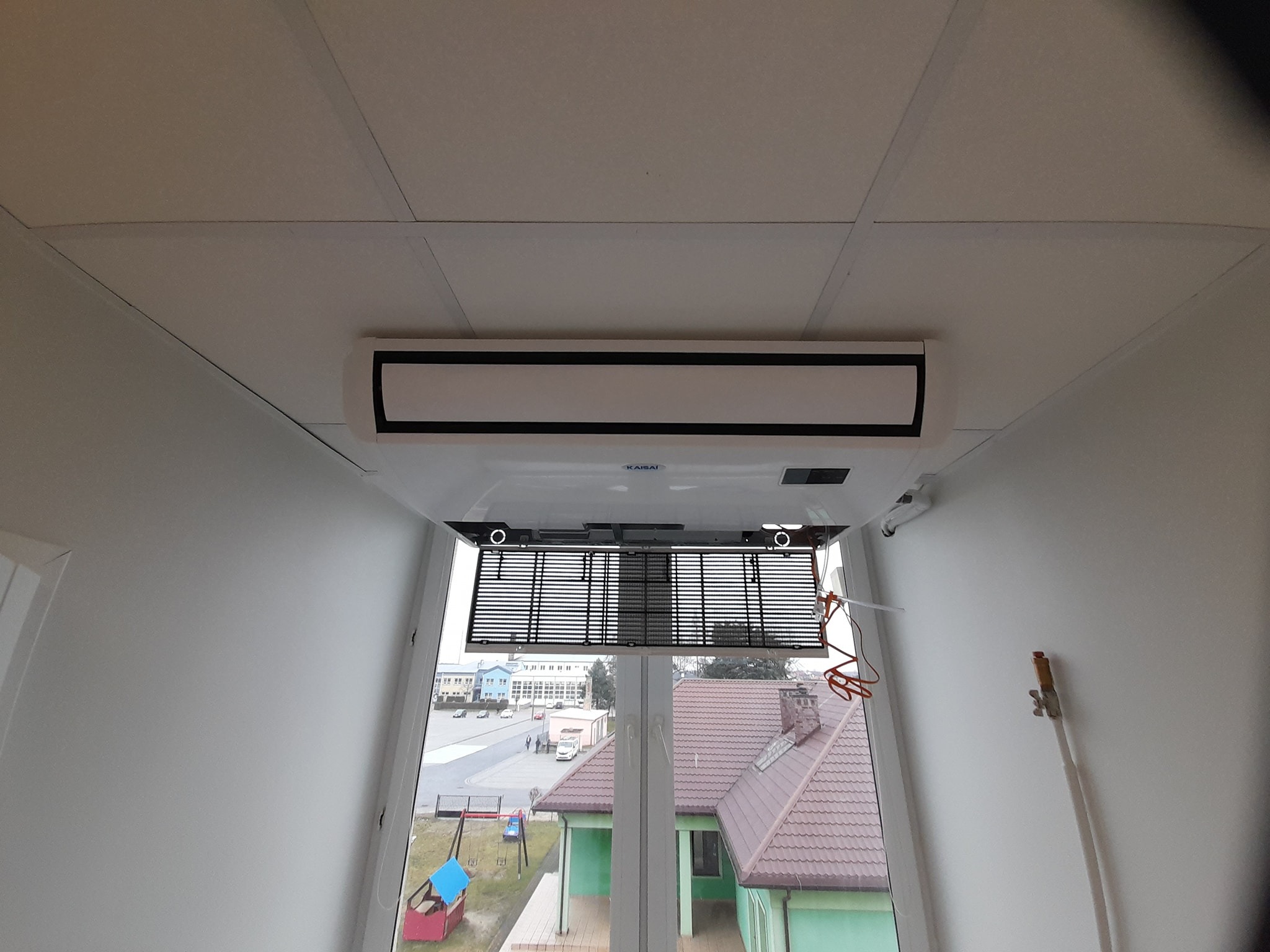 Widok od dołu na zamontowaną kasetonową jednostkę klimatyzacyjną w pomieszczeniu z oknem, widoczne elementy instalacji i budynki na zewnątrz.