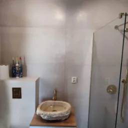 Wykończona łazienka z umywalką z kamienia naturalnego na drewnianym blacie, złota armatura, szklana kabina prysznicowa z deszczownicą i płytki imitujące kamień na ścianach.