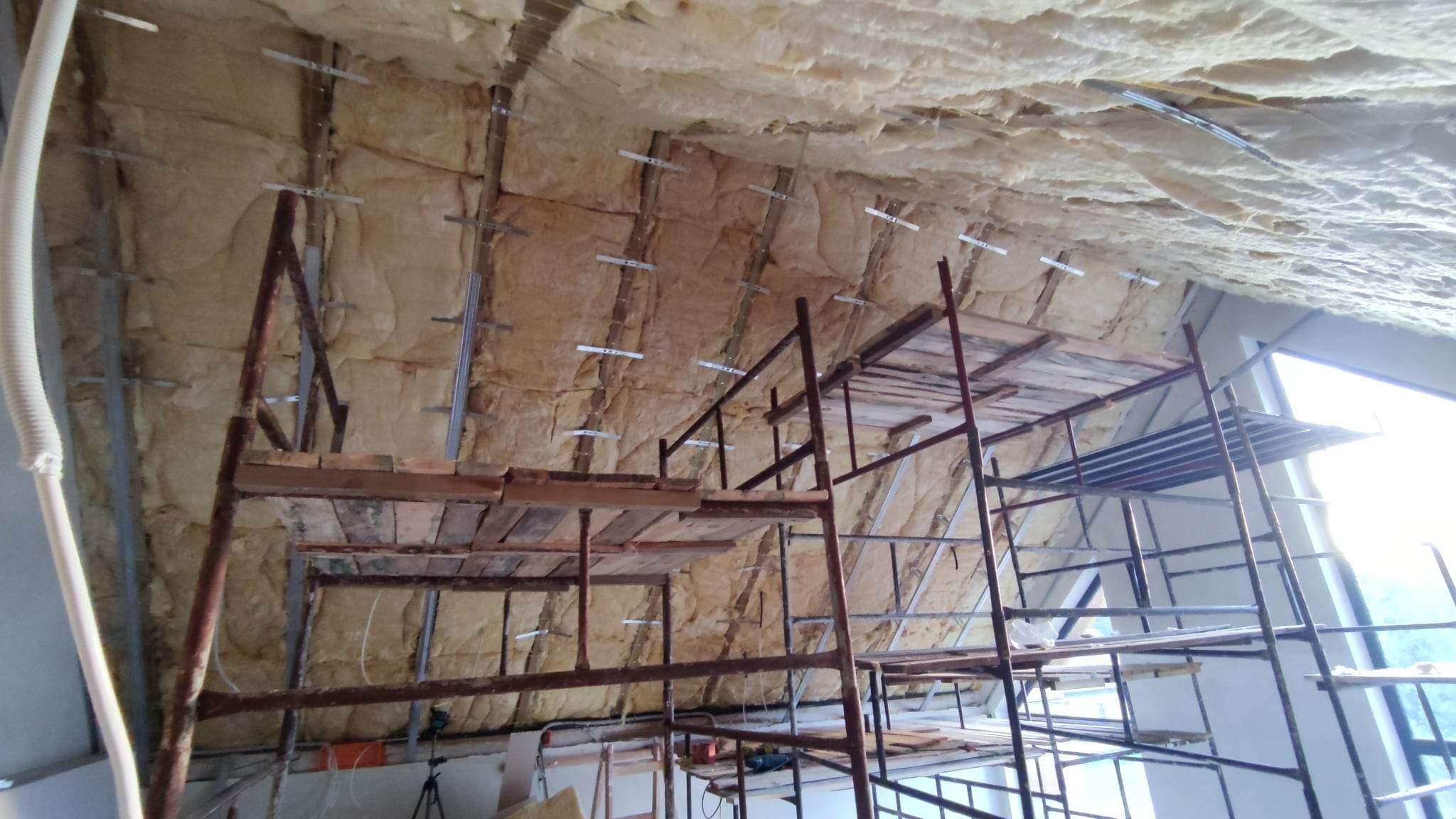 Skośny sufit poddasza w trakcie ocieplania wełną mineralną, widoczne metalowe profile montażowe i rusztowanie z desek, okno w tle.