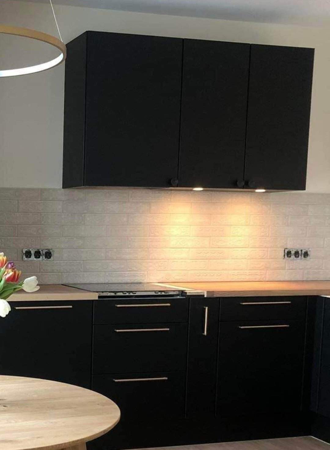 Nowoczesna kuchnia z czarnymi szafkami, drewnianym blatem i białymi płytkami imitującymi cegłę, oświetlona punktowymi lampami podszafkowymi.