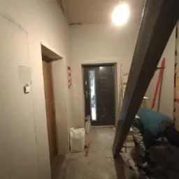 Wnętrze w trakcie remontu z widoczną stalową belką, drabina oparta o ścianę, worki z zaprawą i pracownik w odzieży roboczej