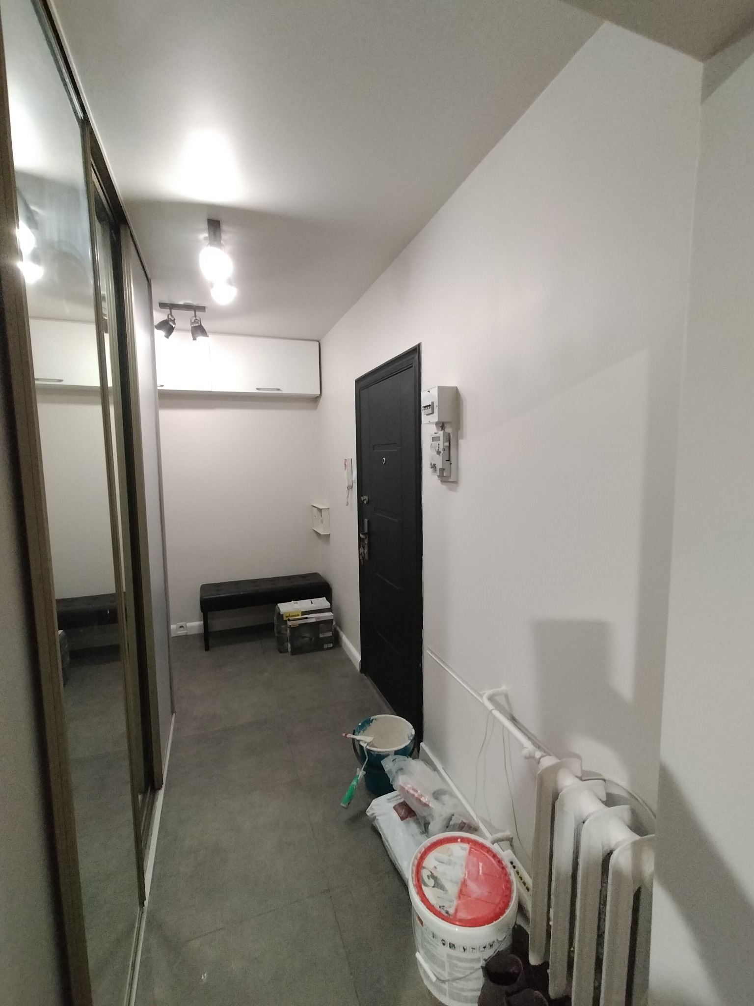 Korytarz w trakcie odświeżania: białe ściany, szara podłoga, widoczne wiadro z farbą, grzejnik i akcesoria malarskie.