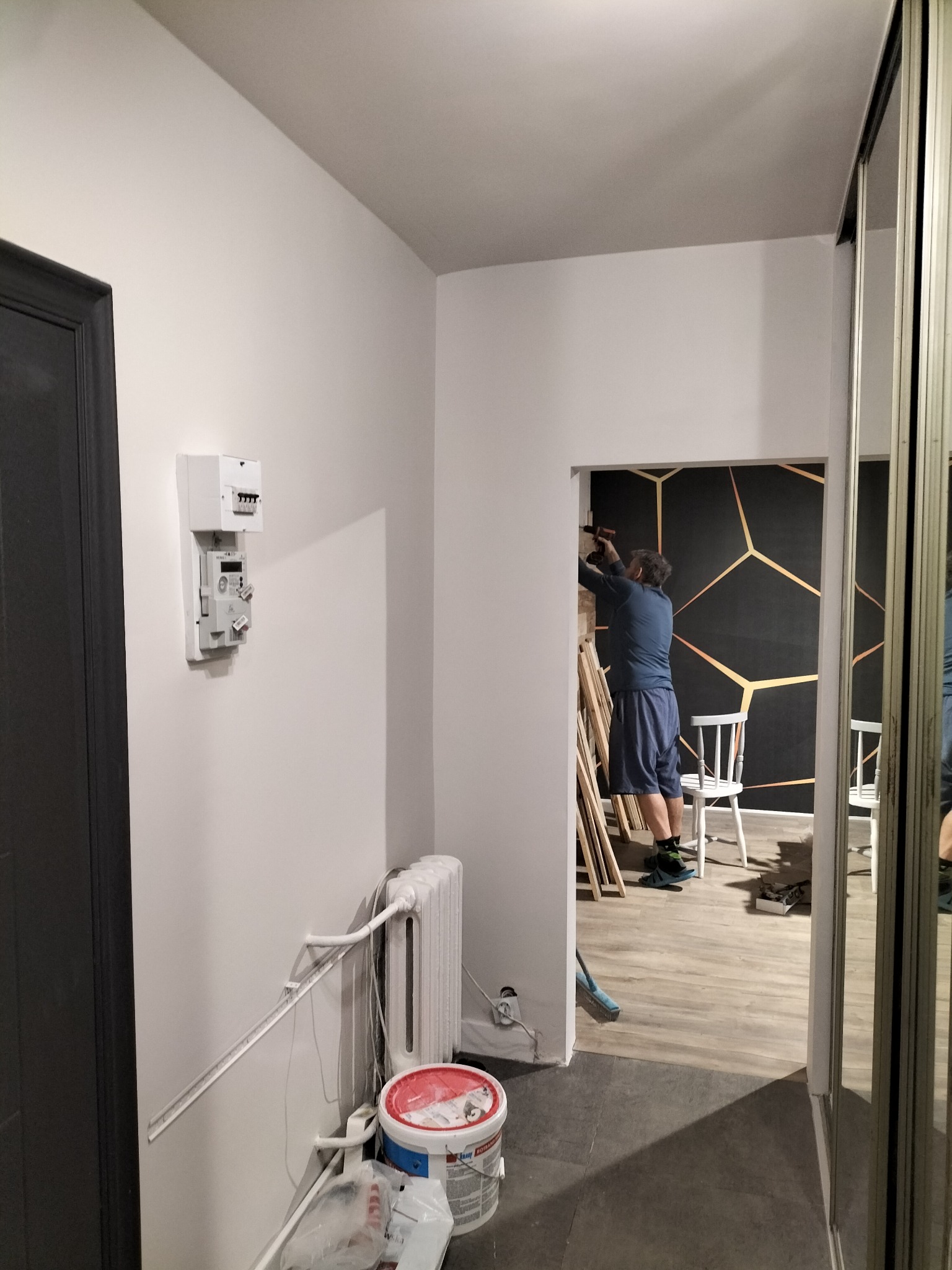 Instalacja dekoracyjnej tapety z geometrycznym wzorem w korytarzu, widoczny pracownik z wiertarką, białe krzesła i niezabezpieczony licznik elektryczny na ścianie.