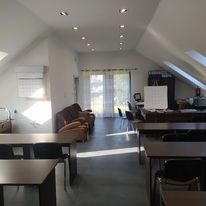 Przestronne, jasne wnętrze gabinetu z poddaszem, widoczne biurka, krzesła, sofa i okna dachowe. Na ścianie kalendarz i tablica suchościeralna. W tle widoczne drzewa za oknem.