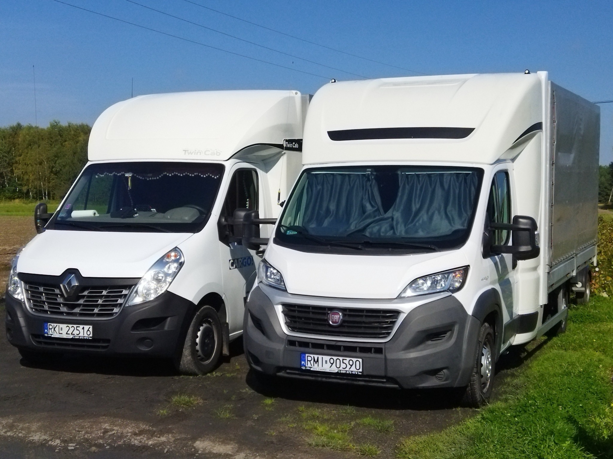 Dwa białe busy dostawcze (Renault Master i Fiat Ducato) z zabudową typu Twin Cab zaparkowane obok siebie na poboczu drogi gruntowej, widoczne tablice rejestracyjne.