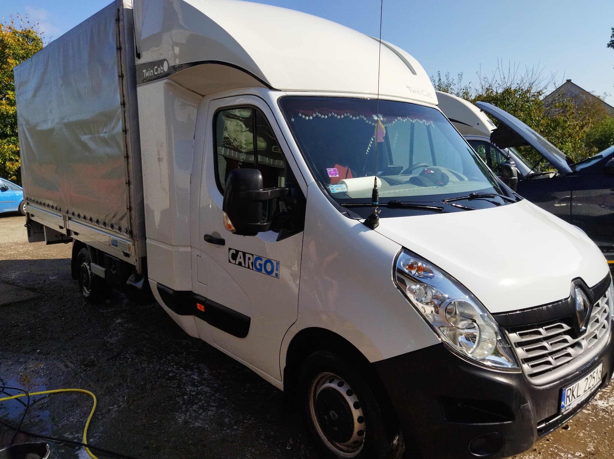 Biały bus typu Twin Cab z plandeką, zaparkowany na zewnątrz z widocznym napisem 'CARGO!' na drzwiach, dekoracyjnymi frędzlami w kabinie i żółtym wężem leżącym na ziemi.