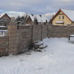 Budowa ogrodzenia z bloczków betonowych w zimowej scenerii, widoczne zbrojenie słupków, paleta z resztkami zaprawy, w tle ośnieżone domy i trampolina.