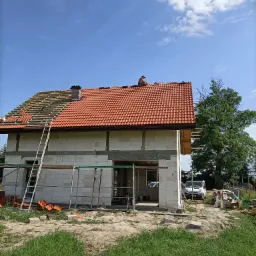 Budowa domu jednorodzinnego z czerwonym dachem w trakcie realizacji, widoczny dekarz na dachu, częściowo ułożona dachówka, rusztowanie i drabina, ściany z białych bloczków.