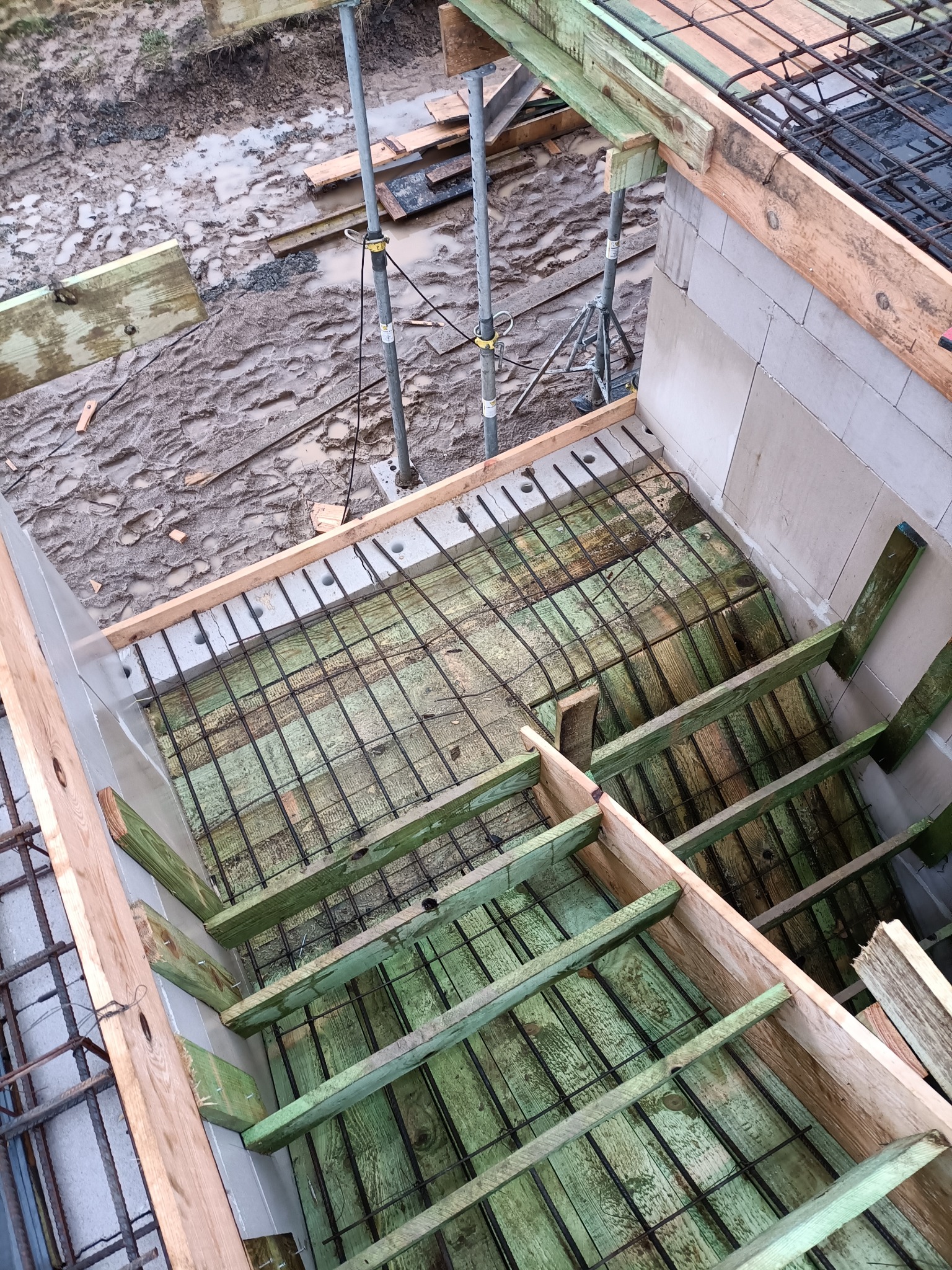 Wiązanie zbrojenia na szalunku pod wylewkę fundamentów w trakcie budowy domu z bloczków betonowych, widok z góry, na zewnątrz błoto po deszczu.