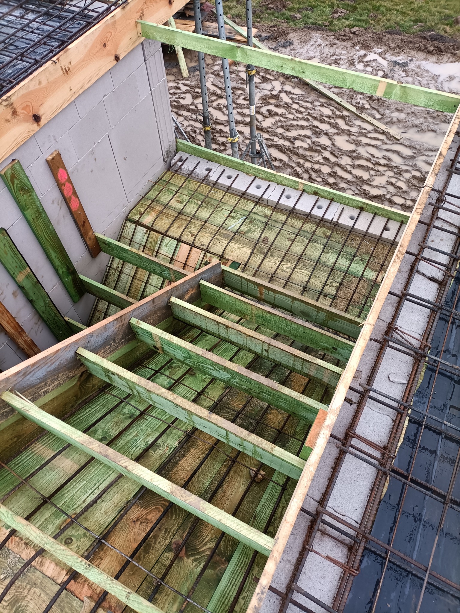 Szczegółowy widok z góry na szalunek z zielonych belek i siatki zbrojeniowej, przygotowany pod wylewkę betonową na budowie domu z bloczków betonowych, z widocznym fragmentem błotnistego terenu w tle.
