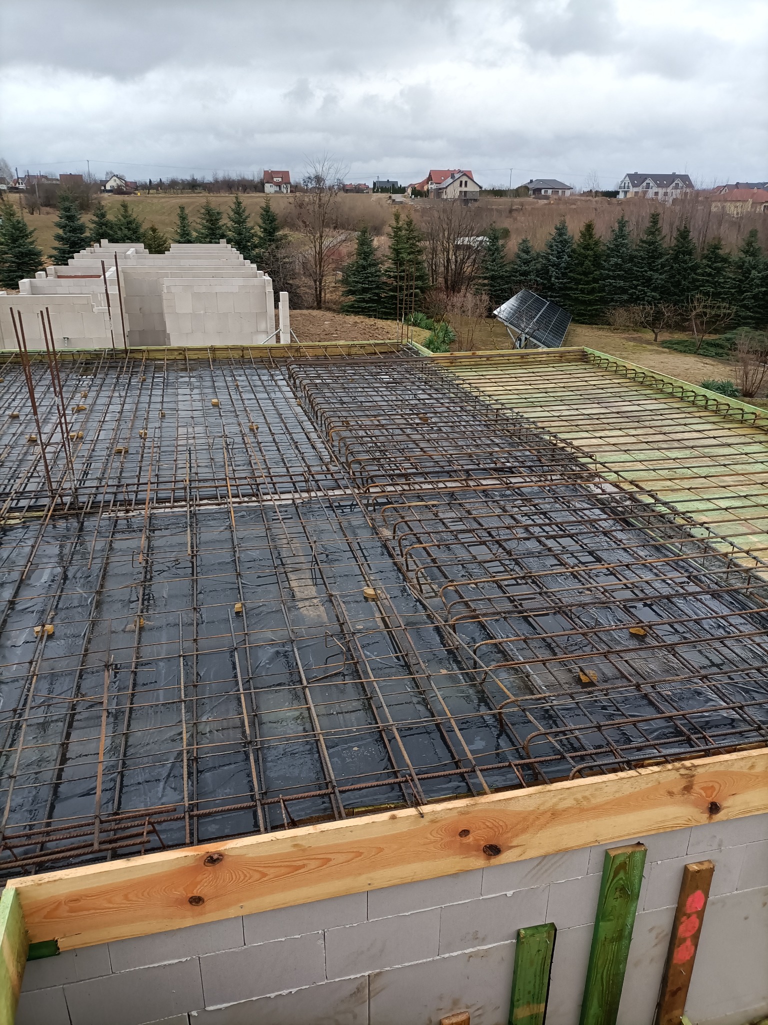 Zbrojenie stropu na budowie domu jednorodzinnego, widoczne siatki z prętów stalowych i szalunek z desek, w tle budynek z pustaków i krajobraz z drzewami i zabudowaniami.