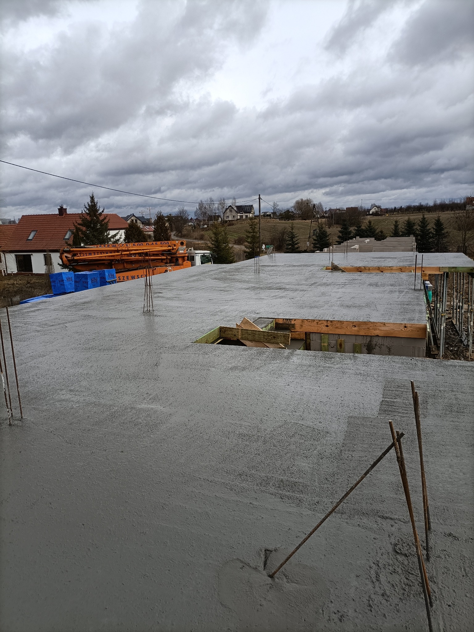 Świeżo wylany beton na stropie domu murowanego z widocznymi wystającymi prętami zbrojeniowymi oraz pomarańczową pompą do betonu w tle, pochmurny dzień.