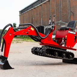 Czerwona, fabrycznie nowa maszyna budowlana na gąsienicach marki Springer, z czarną łyżką i siedziskiem, na tle ceglanego budynku.