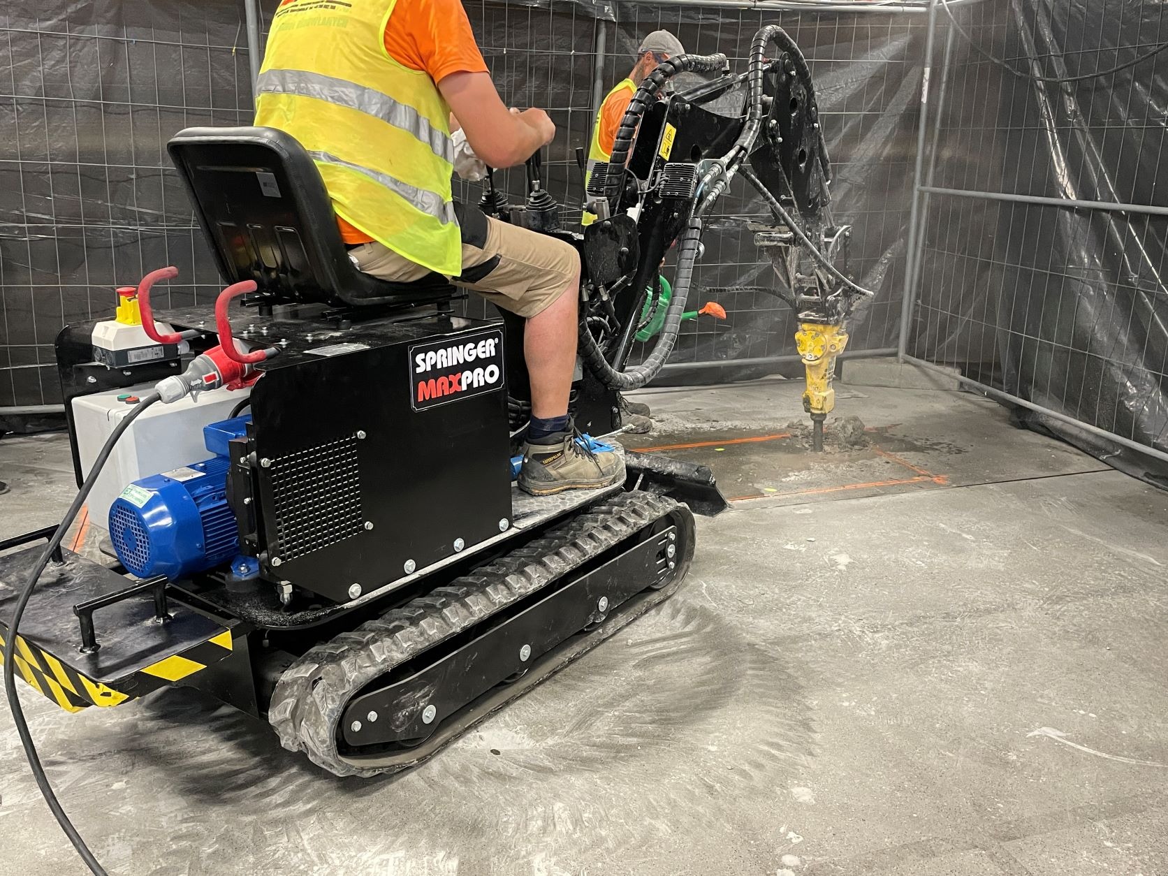 Operator w kamizelce odblaskowej obsługuje małą koparkę Springer Maxpro z młotem hydraulicznym podczas prac wewnątrz budynku. Widoczne ślady po oponach na betonowej podłodze i wyznaczony...