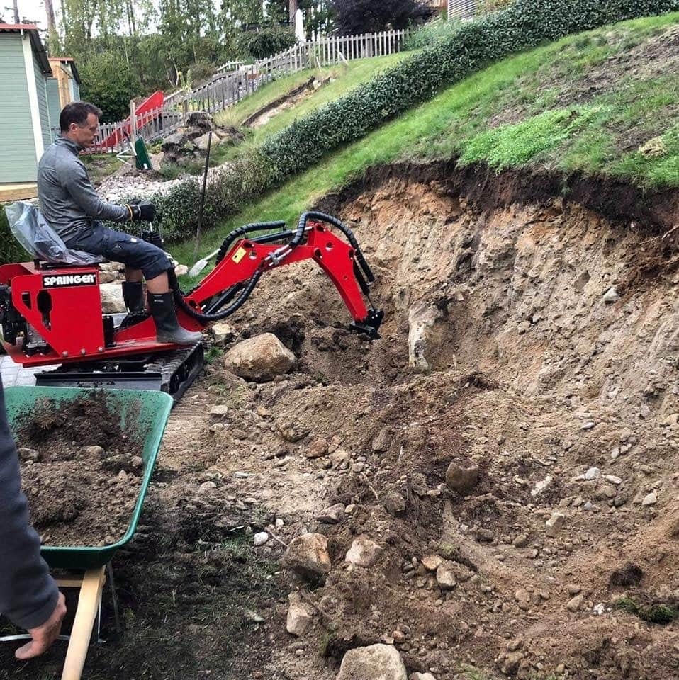 Operator w szarej bluzie steruje czerwoną maszyną budowlaną Springer, wykonując prace ziemne na zboczu wzgórza; w tle widoczny dom i zielony trawnik.