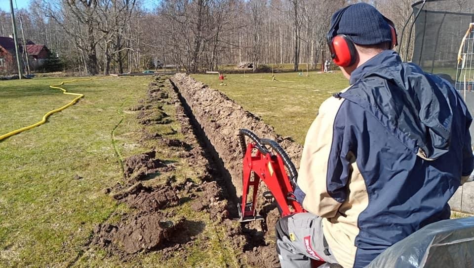 Operator w słuchawkach na minikoparce wykopuje wąski rów na trawniku, widoczny żółty wąż na ziemi.