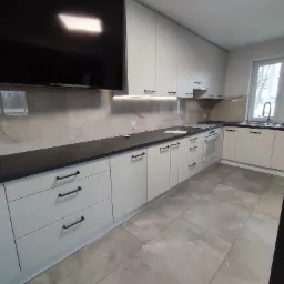 Nowoczesna kuchnia z białymi szafkami, czarnym blatem i marmurowym backsplashiem, oświetlona listwą LED pod szafkami wiszącymi, z widocznym dużym telewizorem na ścianie i oknem z widokiem na zewnątrz.