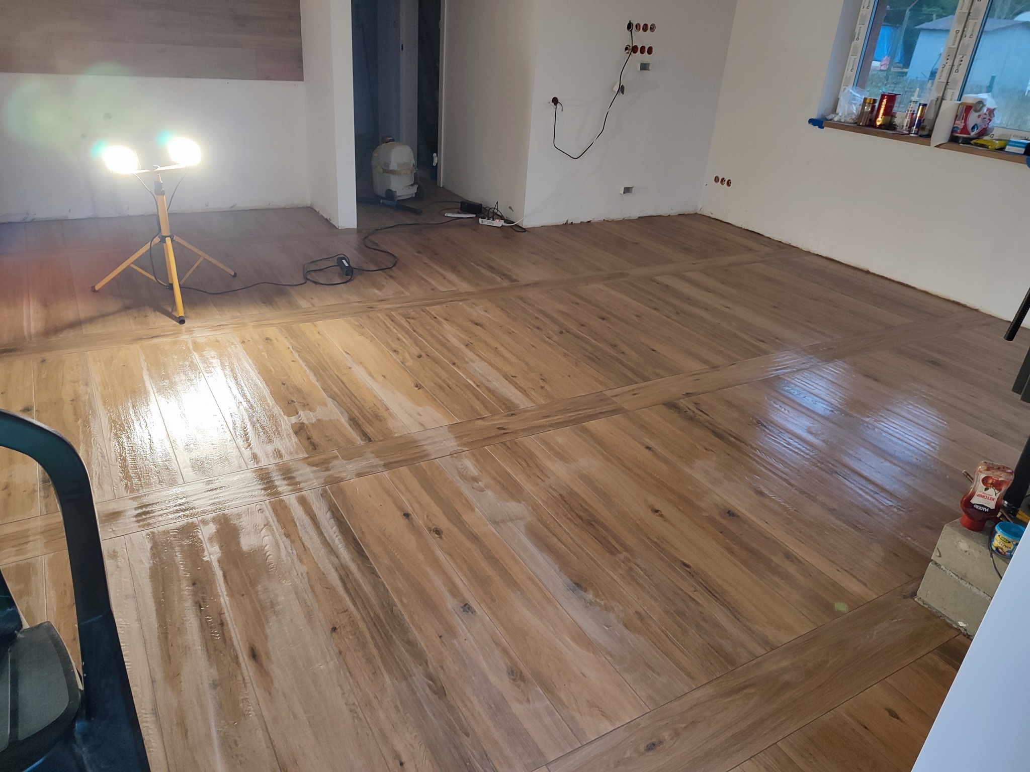 Świeżo położona, lakierowana drewniana podłoga ułożona w jodełkę, oświetlona lampą budowlaną; widoczne puszki elektryczne bez osprzętu i okno z drobnymi przedmiotami na parapecie.