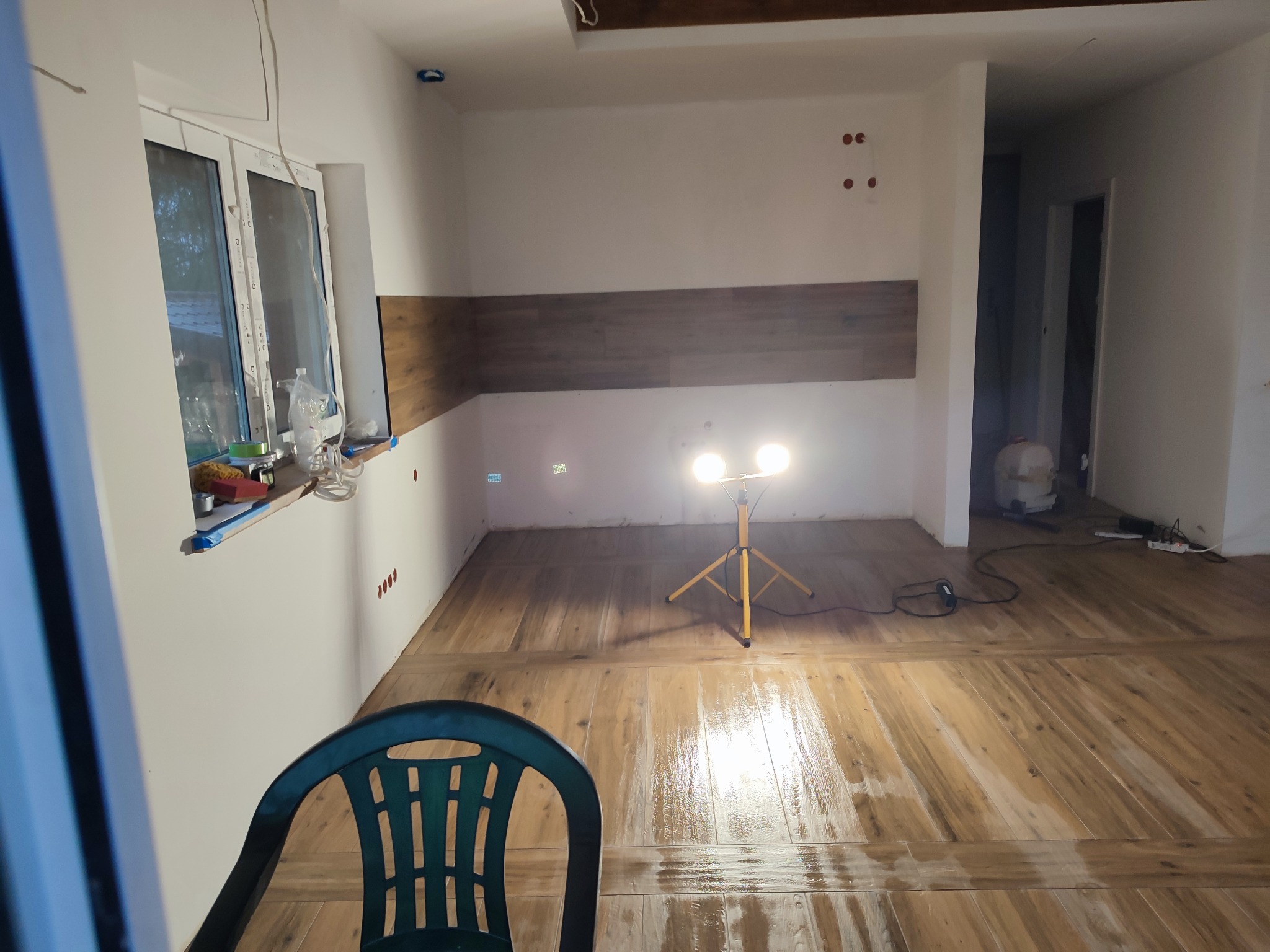 Wnętrze w trakcie prac wykończeniowych: świeżo położona podłoga z płytek imitujących drewno, lampa budowlana na trójnogu oświetla mokrą posadzkę, widoczne ślady po wierceniu w ścianie, plastikowe...