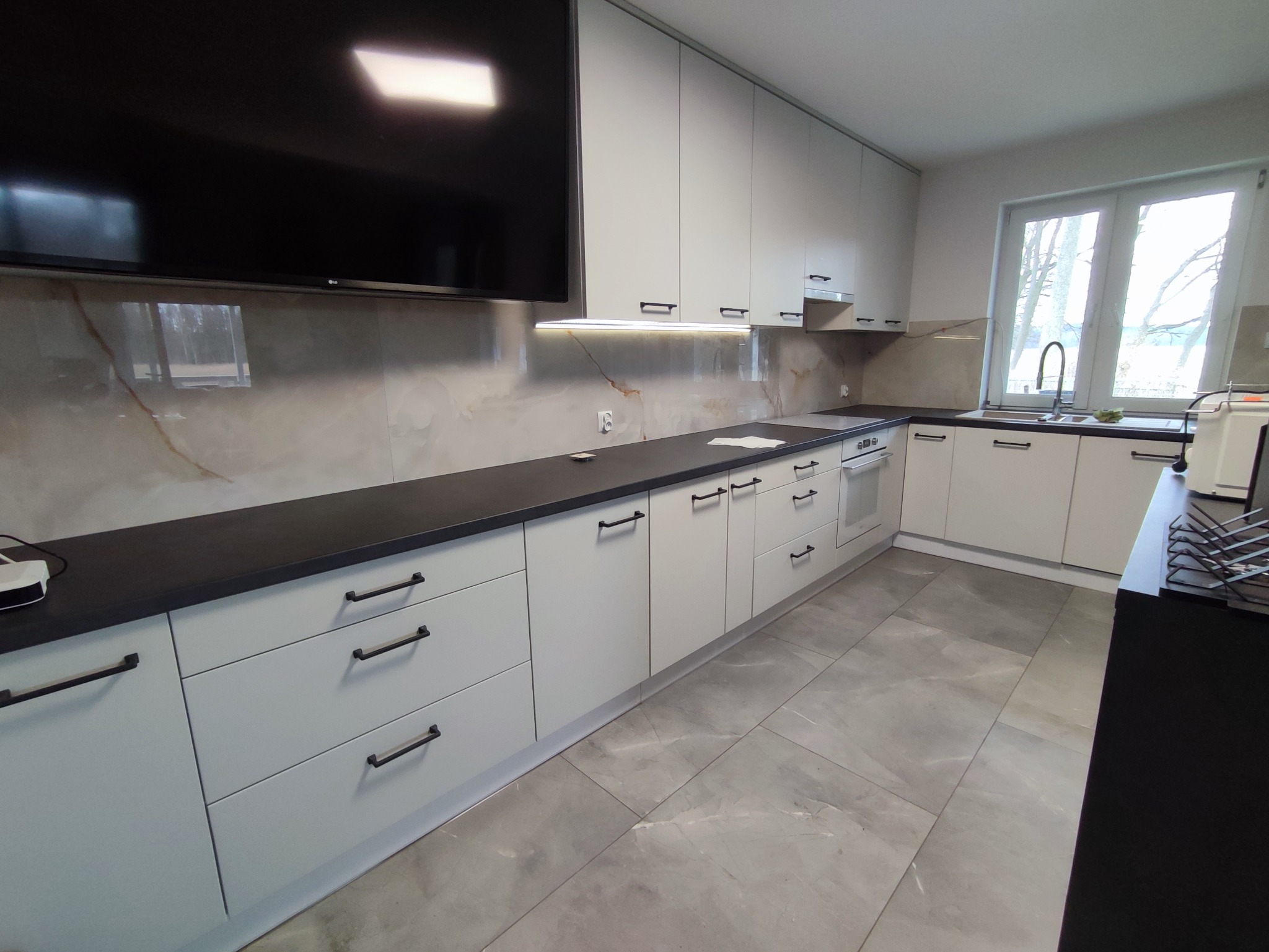 Nowoczesna kuchnia z białymi szafkami, czarnym blatem i marmurowym backsplashiem, oświetlona listwą LED pod szafkami wiszącymi, z widocznym dużym telewizorem na ścianie i oknem z widokiem na zewnątrz.