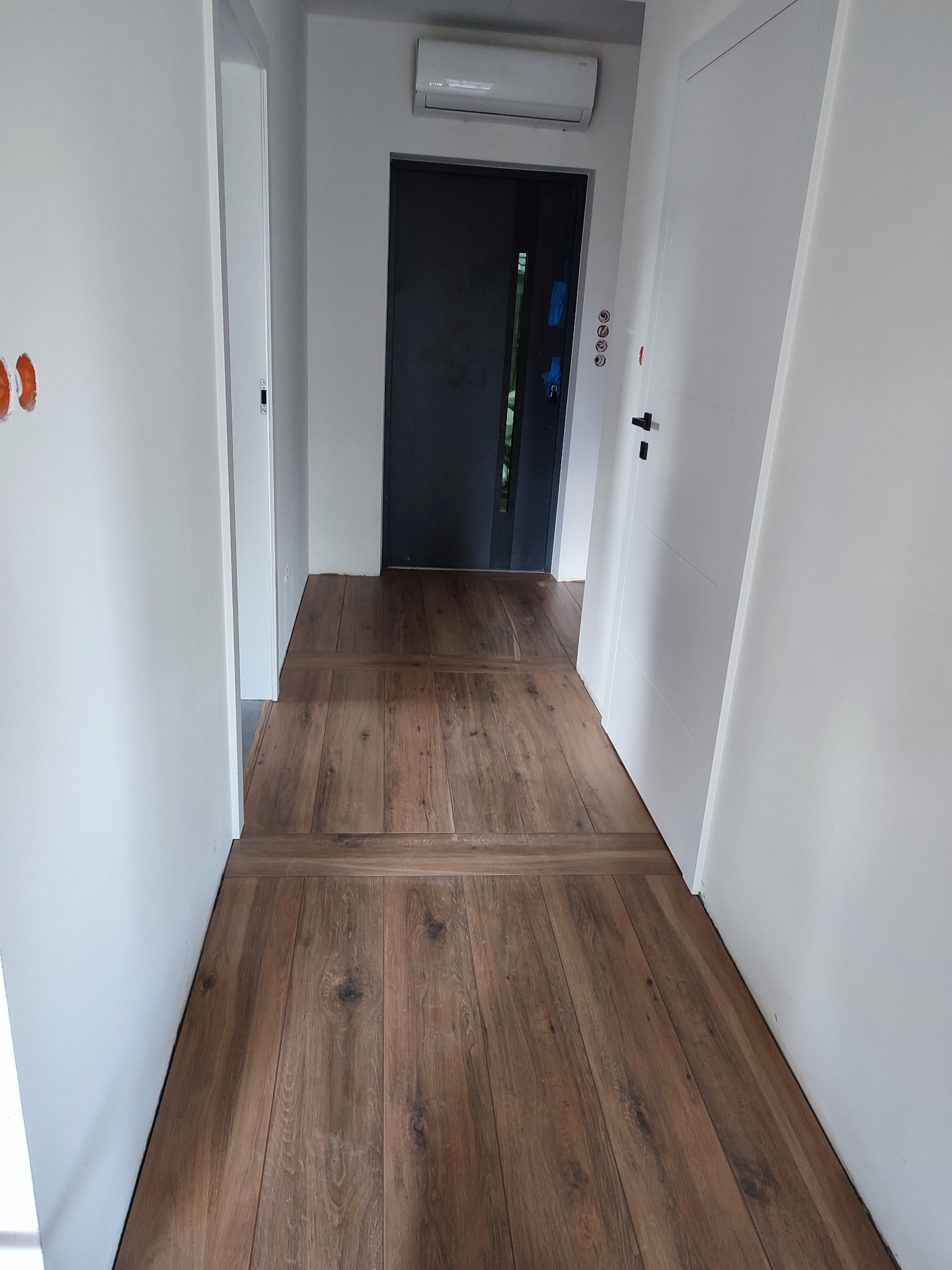 Korytarz z nową, drewnopodobną podłogą ułożoną w pasy, białe ściany z widocznymi puszkami elektrycznymi, białe drzwi wewnętrzne i ciemne drzwi wejściowe.