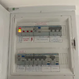 Otwarta skrzynka elektryczna z widocznymi bezpiecznikami, wyłącznikiem głównym, kontrolkami faz i opisami obwodów, w tym płyty indukcyjnej i oświetlenia.