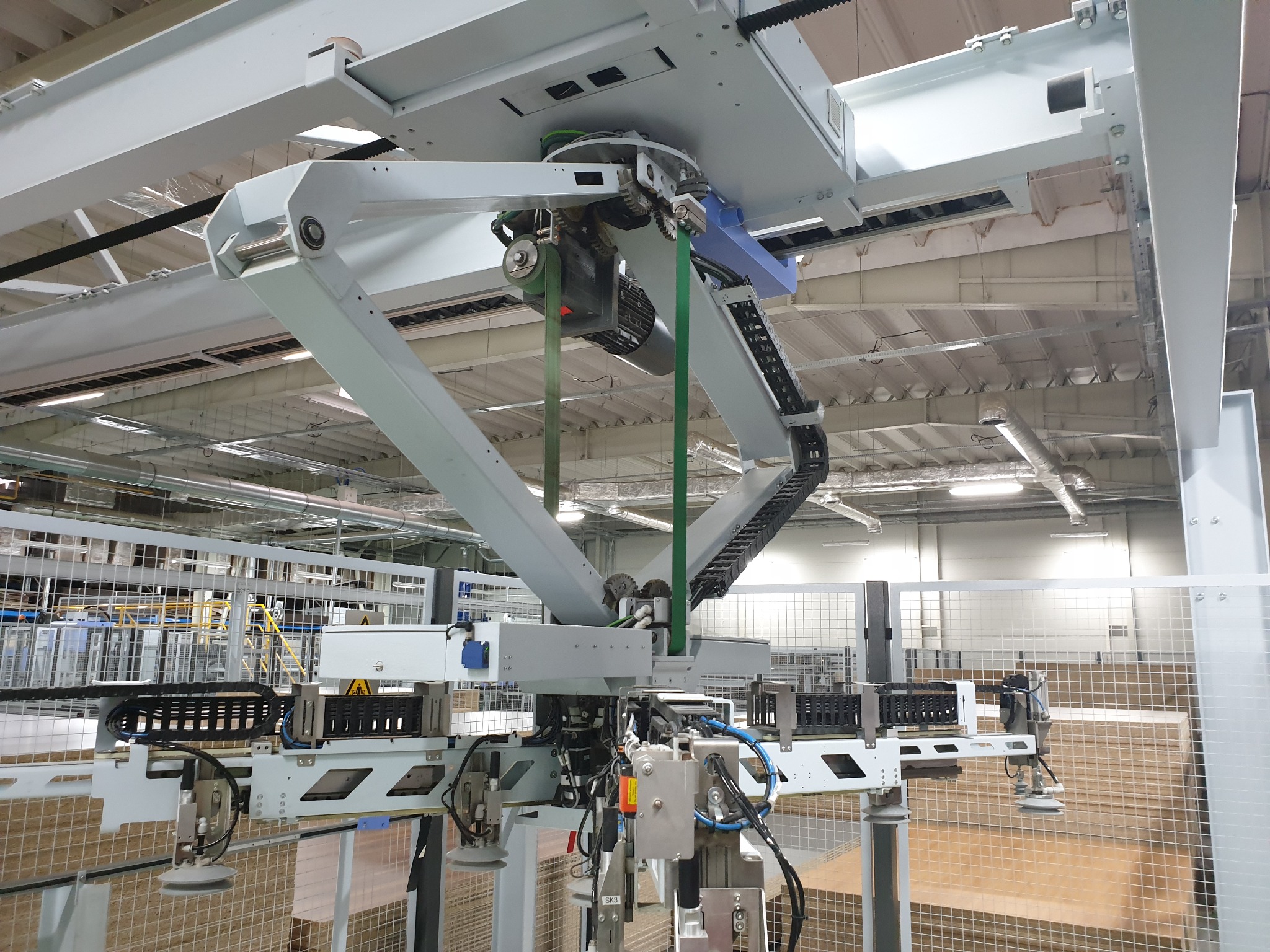 Automatyczna linia produkcyjna z robotem przemysłowym podnoszącym płyty w hali produkcyjnej, widoczne elementy mechaniczne, silniki, kable i czujniki.