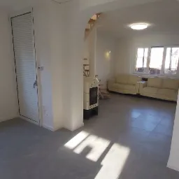 Wyremontowane wnętrze z szarymi płytkami podłogowymi imitującymi marmur, białe ściany, grzejnik kaflowy i widoczne schody w głębi, oświetlone promieniami słońca wpadającymi przez okno.