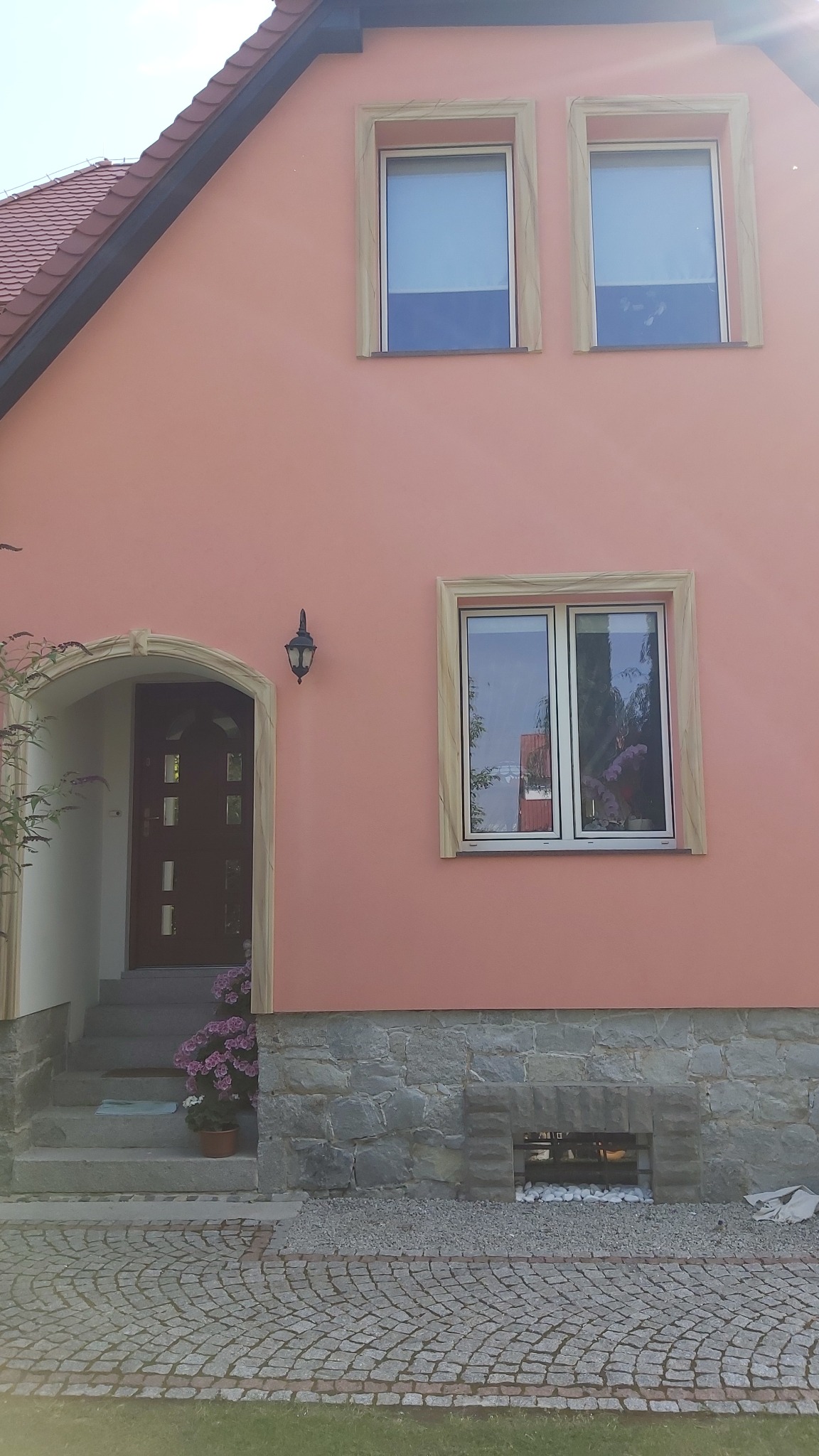 Różowa elewacja domu jednorodzinnego z kamienną podmurówką, ozdobnymi obramowaniami okien i wejścia, oraz brukowanym podjazdem.