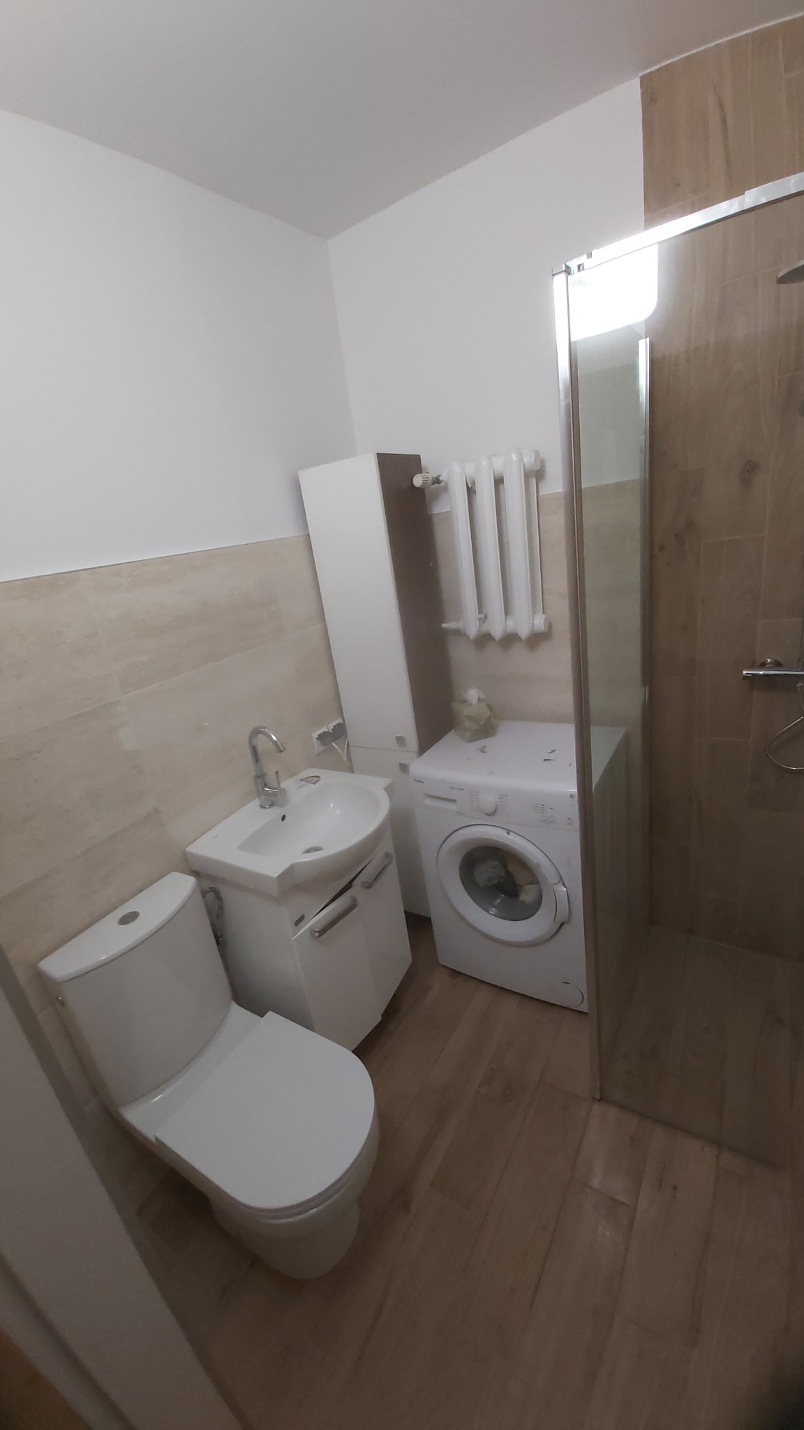 Kompletna biała łazienka z umywalką, toaletą, pralką i kabiną prysznicową, wykończona płytkami w odcieniach beżu i brązu, z grzejnikiem na ścianie i podłogą imitującą drewno.
