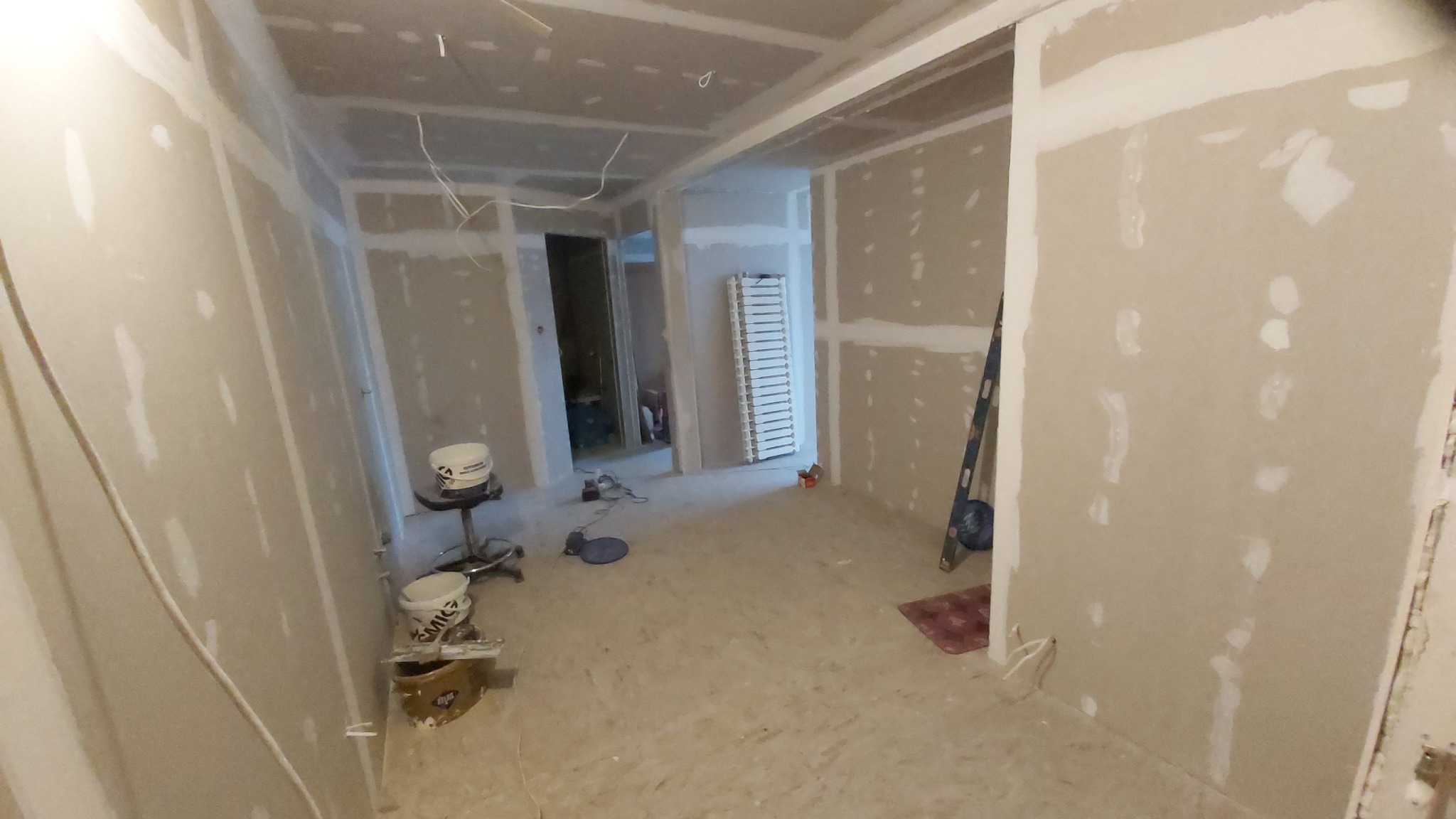 Wnętrze w trakcie prac wykończeniowych: ściany z płyt gipsowo-kartonowych z widocznymi łączeniami, szpachla, wiadro z farbą na stołku warsztatowym, narzędzia malarskie i poziomica oparta o ścianę.