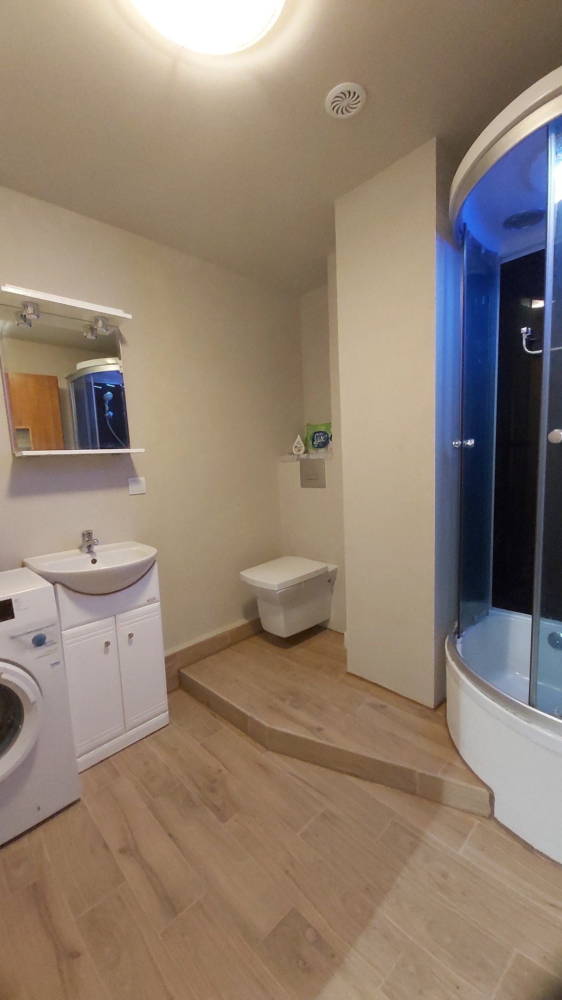 Jasna łazienka z drewnopodobnymi płytkami, kabiną prysznicową z niebieskim podświetleniem, umywalką z szafką, pralką i podwieszaną toaletą na podwyższeniu.