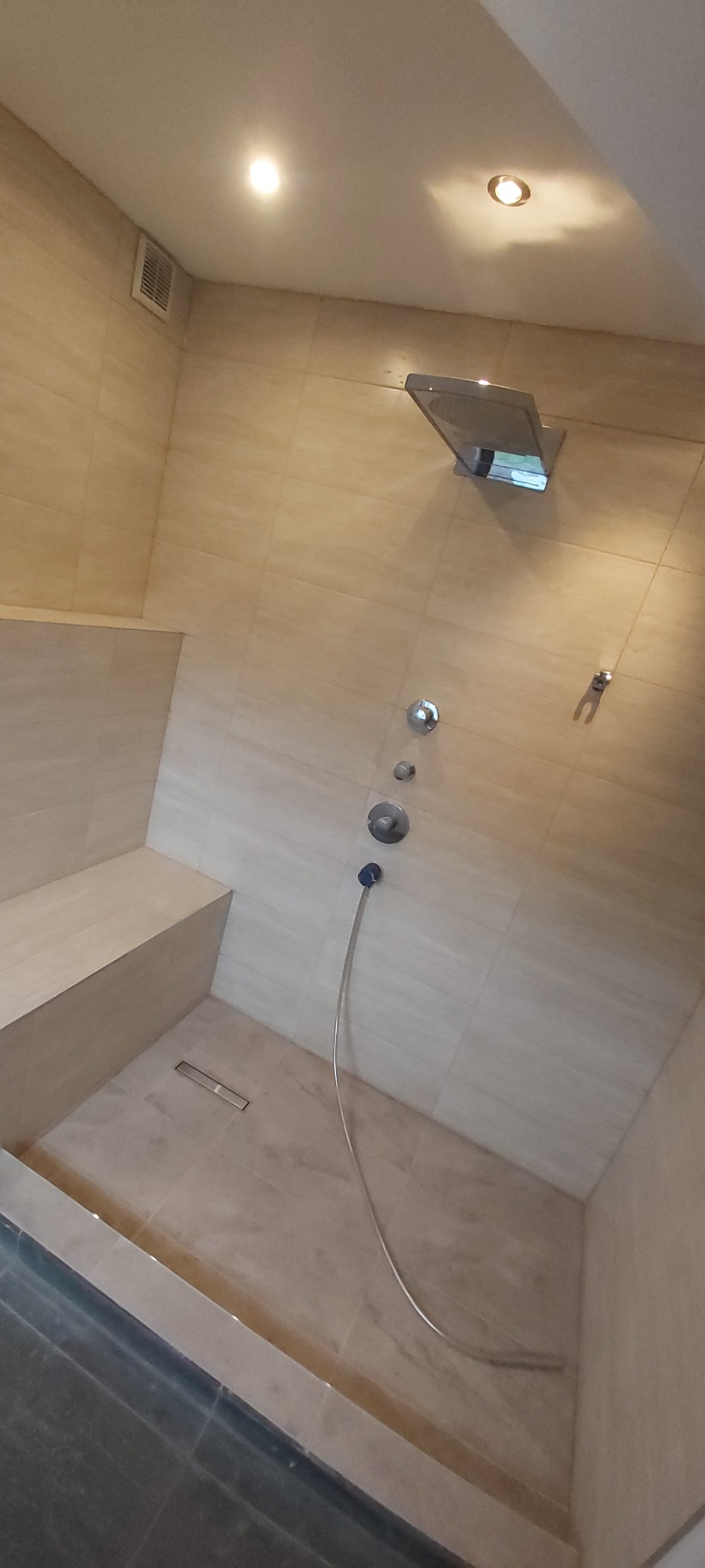 Wykończona kabina prysznicowa z siedziskiem wyłożona jasnymi płytkami imitującymi trawertyn, z deszczownicą i odpływem liniowym w podłodze.
