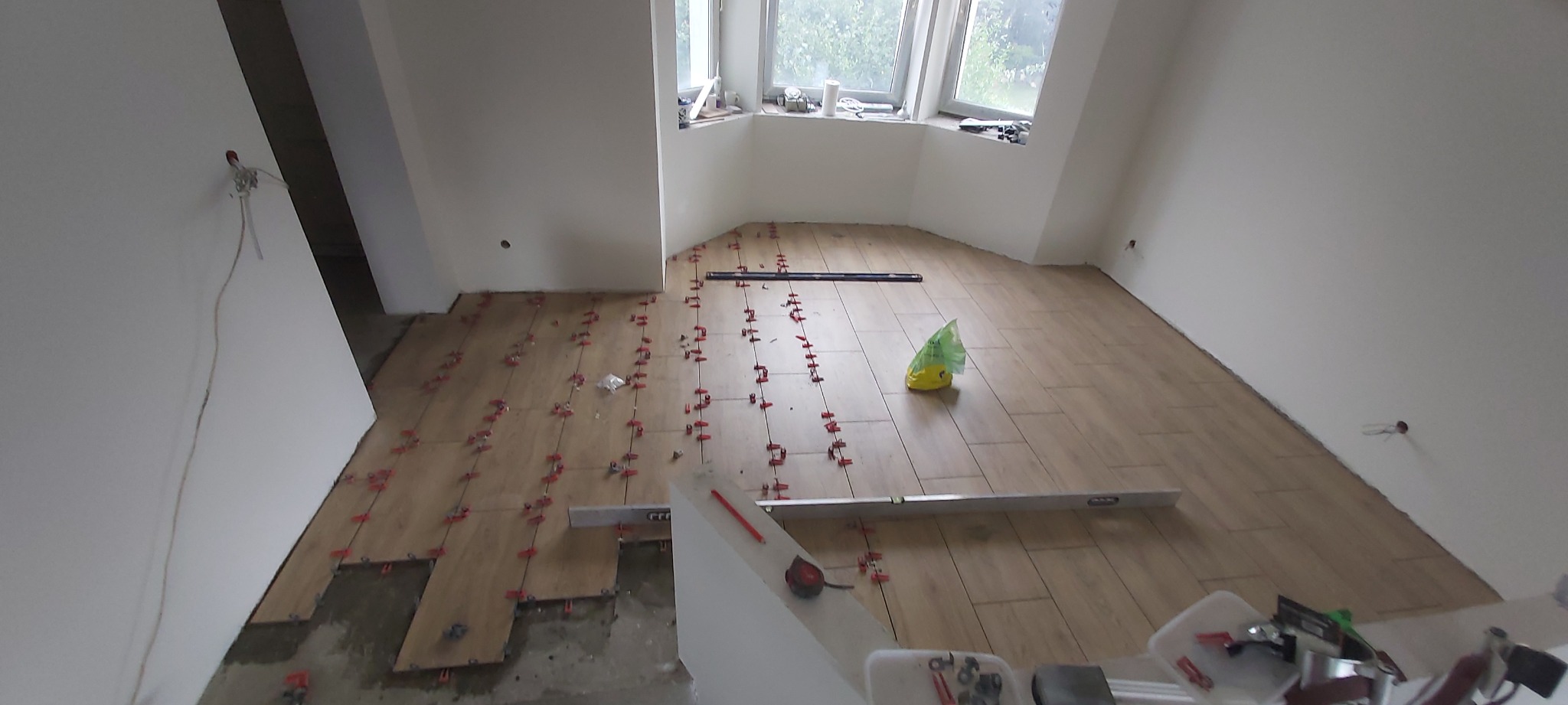 Układanie podłogi z płytek imitujących drewno w jasnym pomieszczeniu z oknem wykuszowym, widoczne poziomice i system poziomowania płytek.