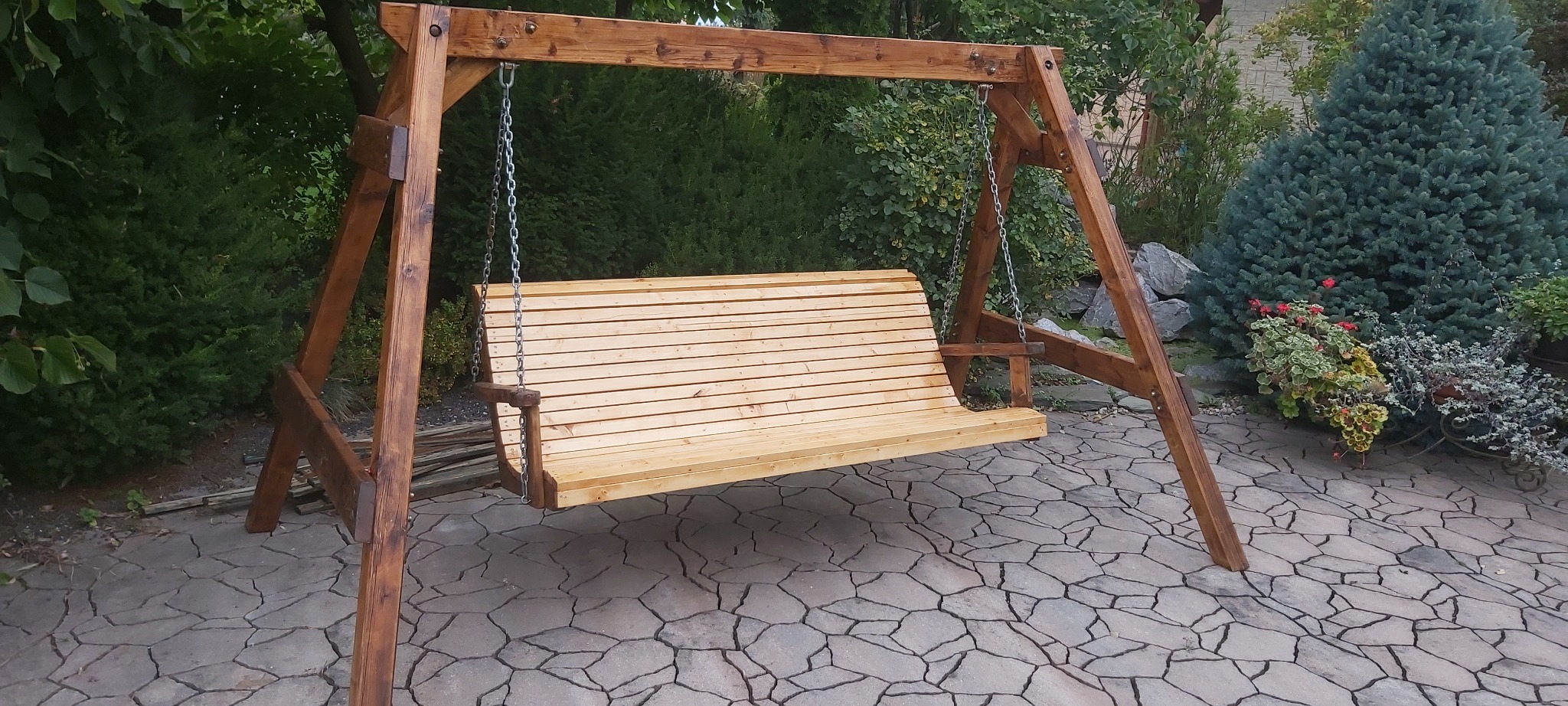 Drewniana huśtawka ogrodowa z jasnego drewna na łańcuchach, z ciemnobrązową konstrukcją, ustawiona na kamiennej posadzce w ogrodzie w Legnicy.