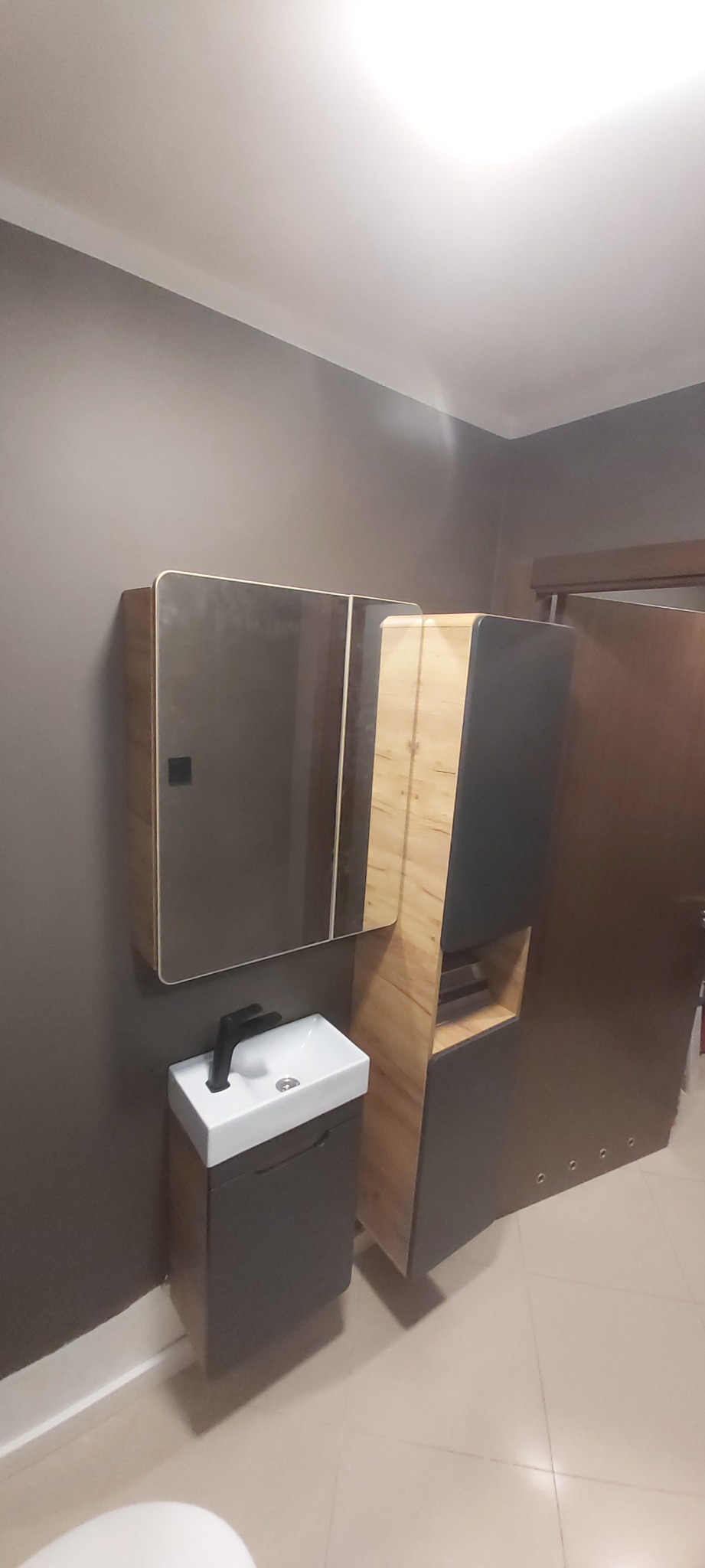 Nowoczesna łazienka z szarą ścianą, wiszącą szafką z umywalką, lustrem i wysoką szafką w kolorze drewna i szarości.