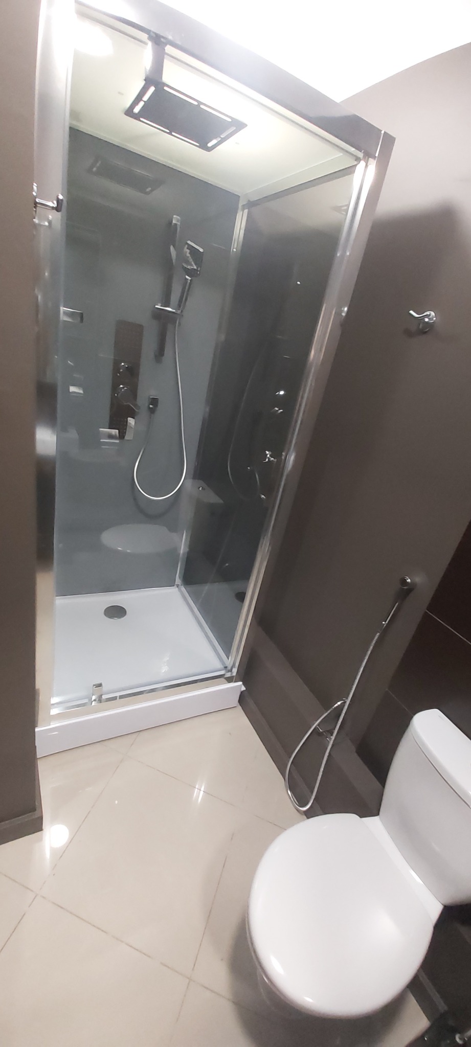 Nowoczesna kabina prysznicowa z deszczownicą i hydromasażem w łazience z ciemnymi ścianami i beżowymi płytkami podłogowymi, widoczna toaleta z bidetką.