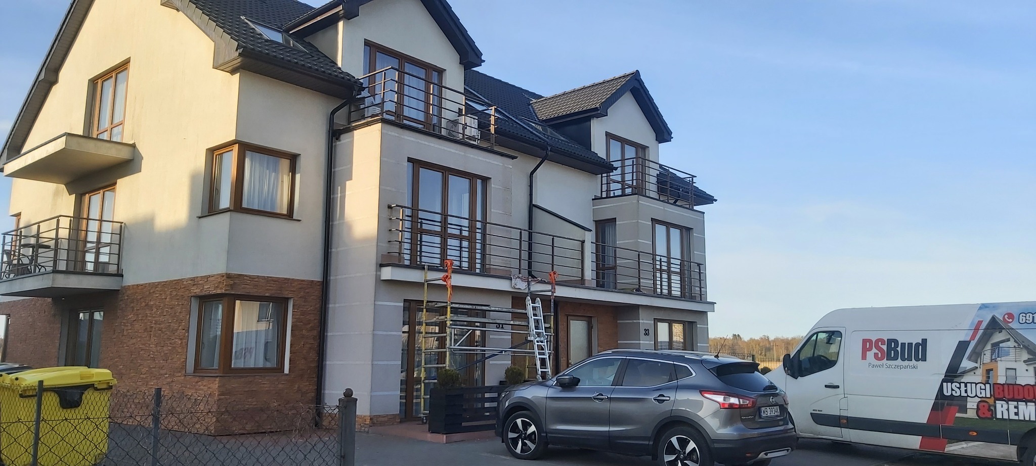 Dwupiętrowy dom z balkonami w trakcie prac wykończeniowych elewacji, widoczne rusztowania i drabina, obok zaparkowany samochód i bus firmy budowlanej z logo.