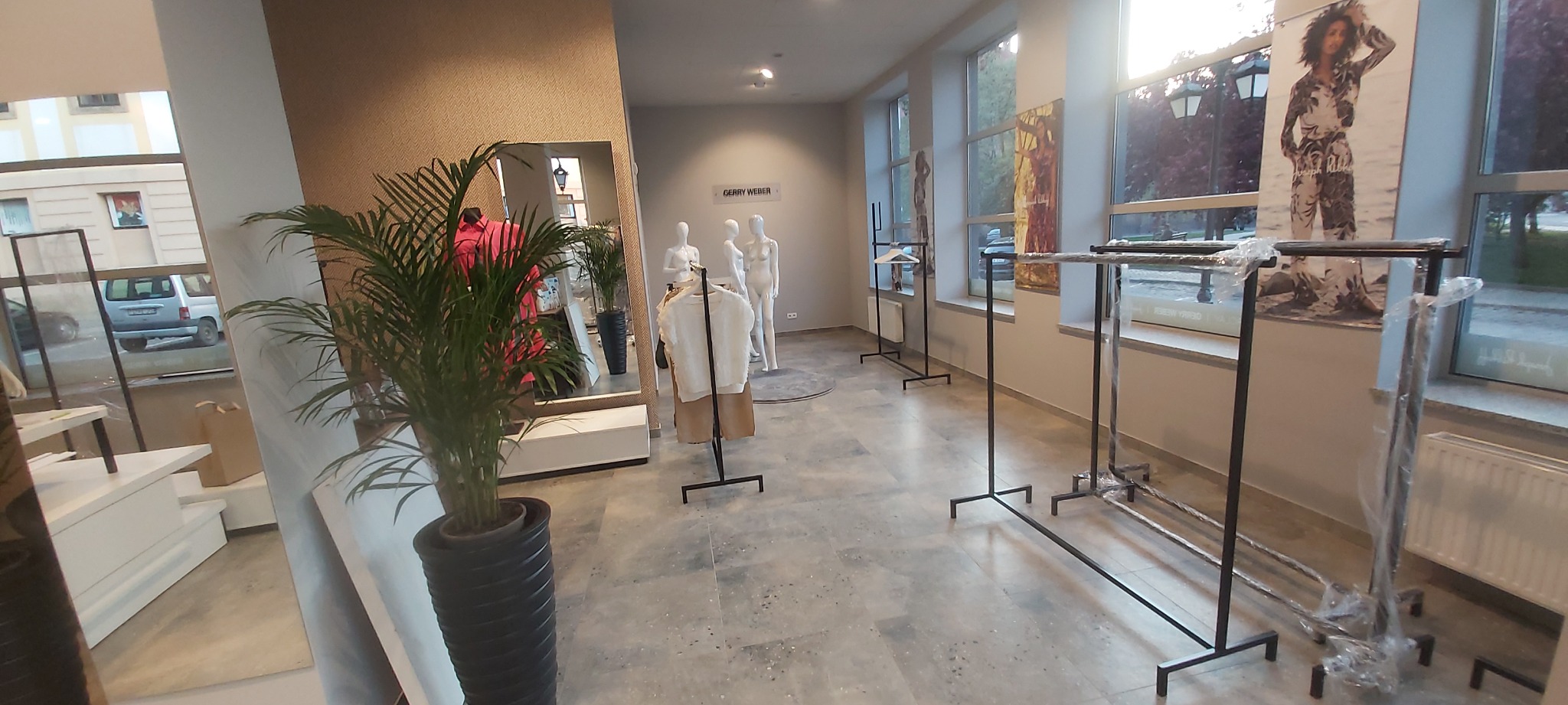 Wnętrze sklepu odzieżowego z manekinami, wieszakami i rośliną doniczkową w czarnej donicy. W tle widoczne okna z plakatami i zabudowa sklepu.