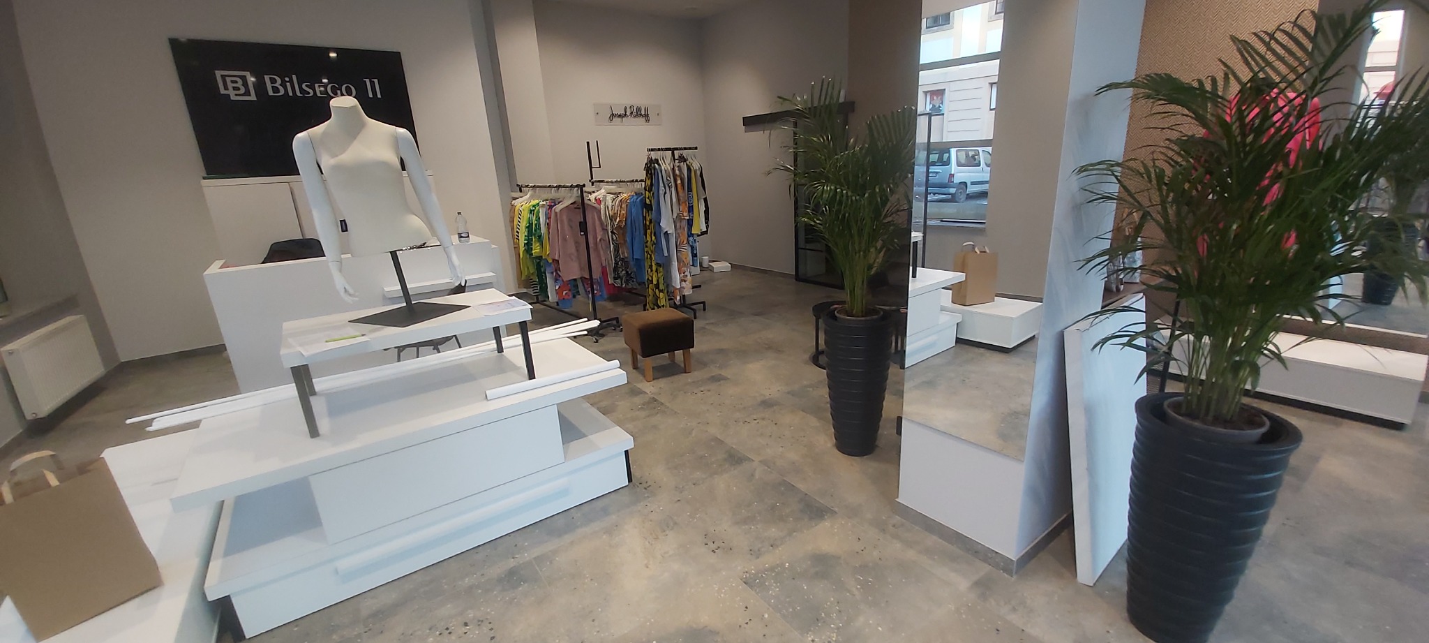 Wnętrze sklepu odzieżowego z manekinem bez głowy na pierwszym planie, w tle wieszaki z ubraniami i duże donice z roślinami.