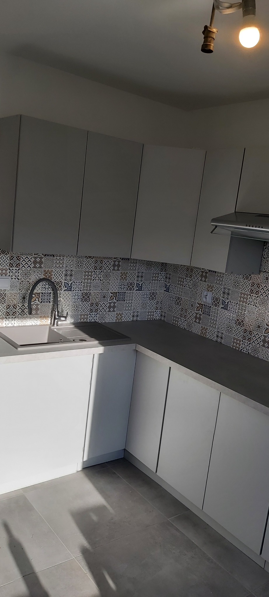 Jasna kuchnia z białymi szafkami i blatem w kolorze szarości, ozdobiona mozaiką na ścianie nad blatem, widoczny zlew i bateria, z lampą sufitową w górnym rogu.