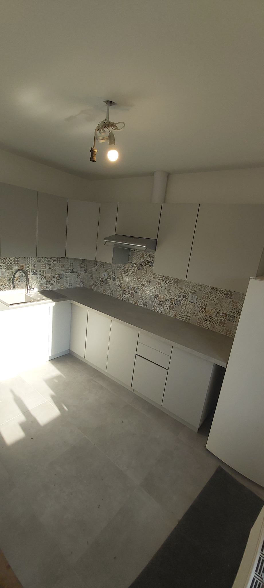 Jasna kuchnia z białymi szafkami, szarym blatem i mozaiką nad blatem. W tle widoczna lampa z żarówką na kablu.