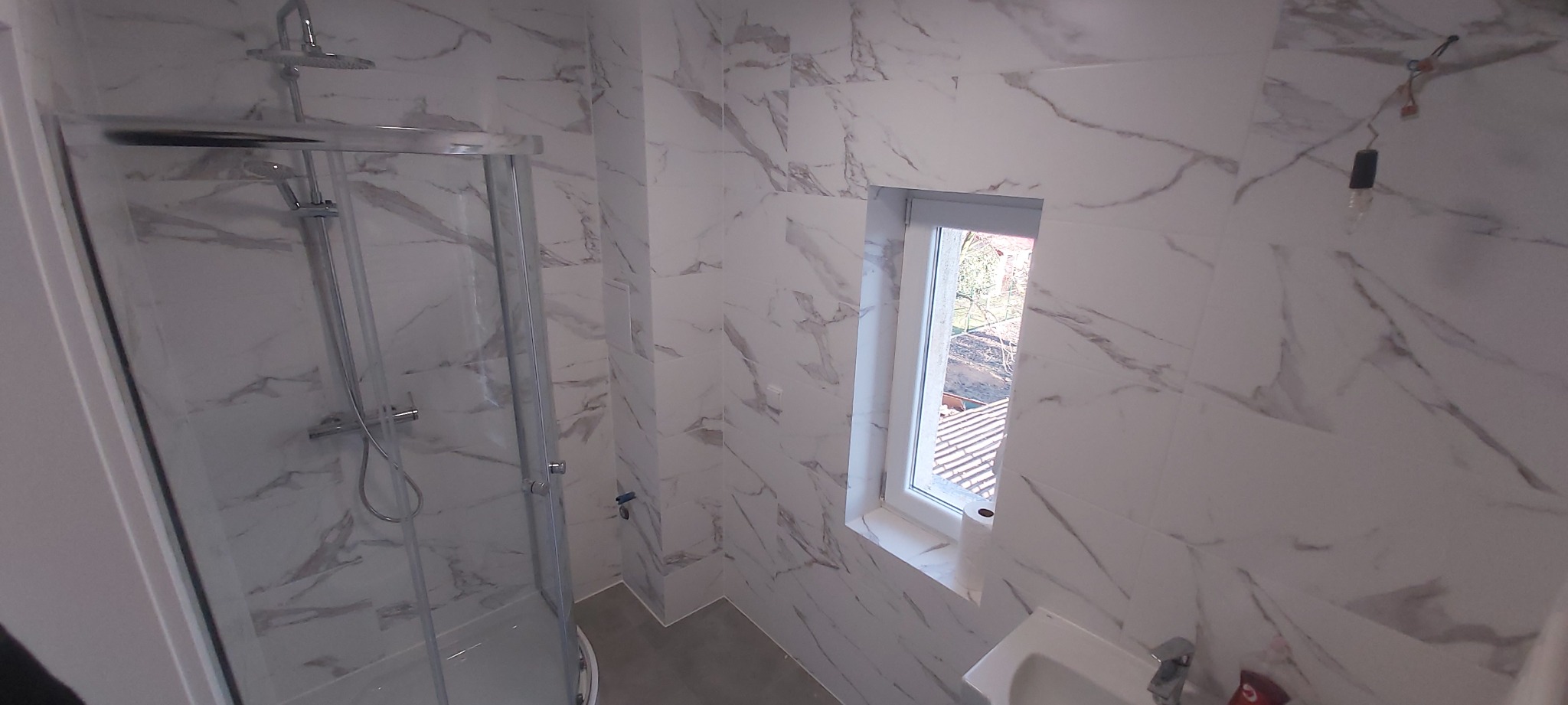 Nowoczesna łazienka z kabiną prysznicową i marmurowymi płytkami na ścianach, małe okno z widokiem na dach budynku, minimalistyczna umywalka.