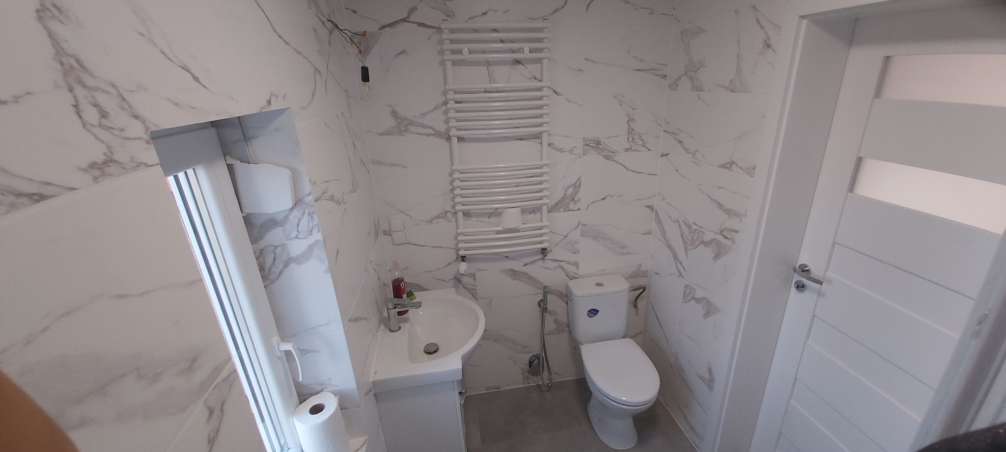 Wykończona łazienka z marmurowymi płytkami na ścianach, białą umywalką, toaletą i grzejnikiem drabinkowym, widoczne wystające kable elektryczne przy suficie.