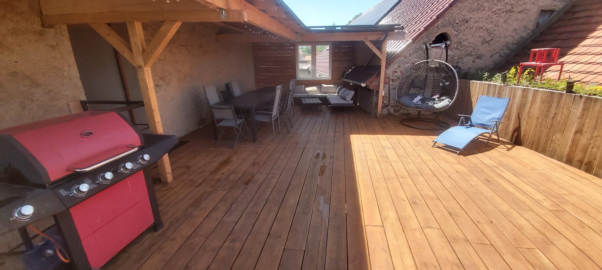 Drewniany taras zadaszony częściowo drewnianą konstrukcją, wyposażony w meble ogrodowe: stół z krzesłami, kanapę, wiszący fotel i leżak, z czerwonym grillem na pierwszym planie.