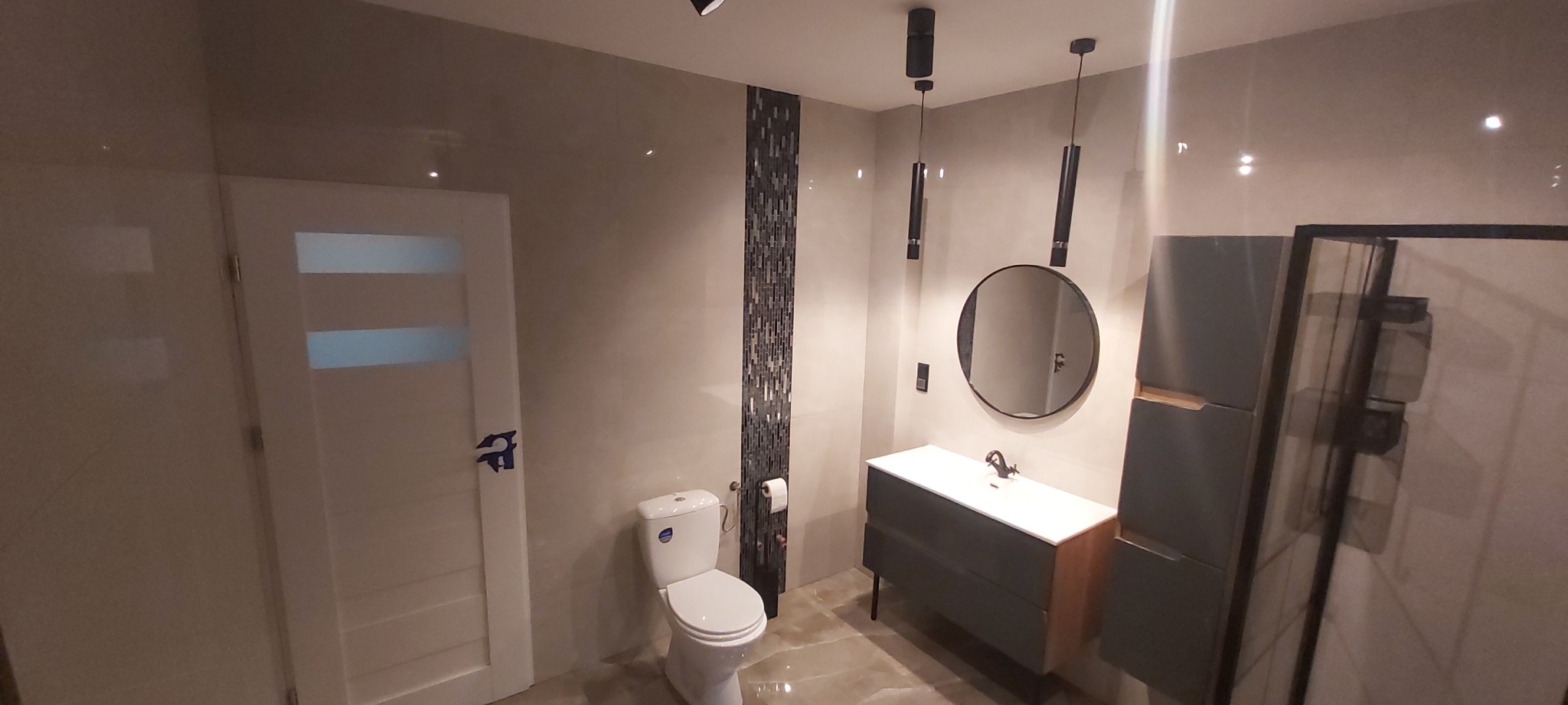 Nowoczesna łazienka z beżowymi płytkami, czarnymi lampami wiszącymi i okrągłym lustrem nad umywalką z szafką; widoczna toaleta i kabina prysznicowa.